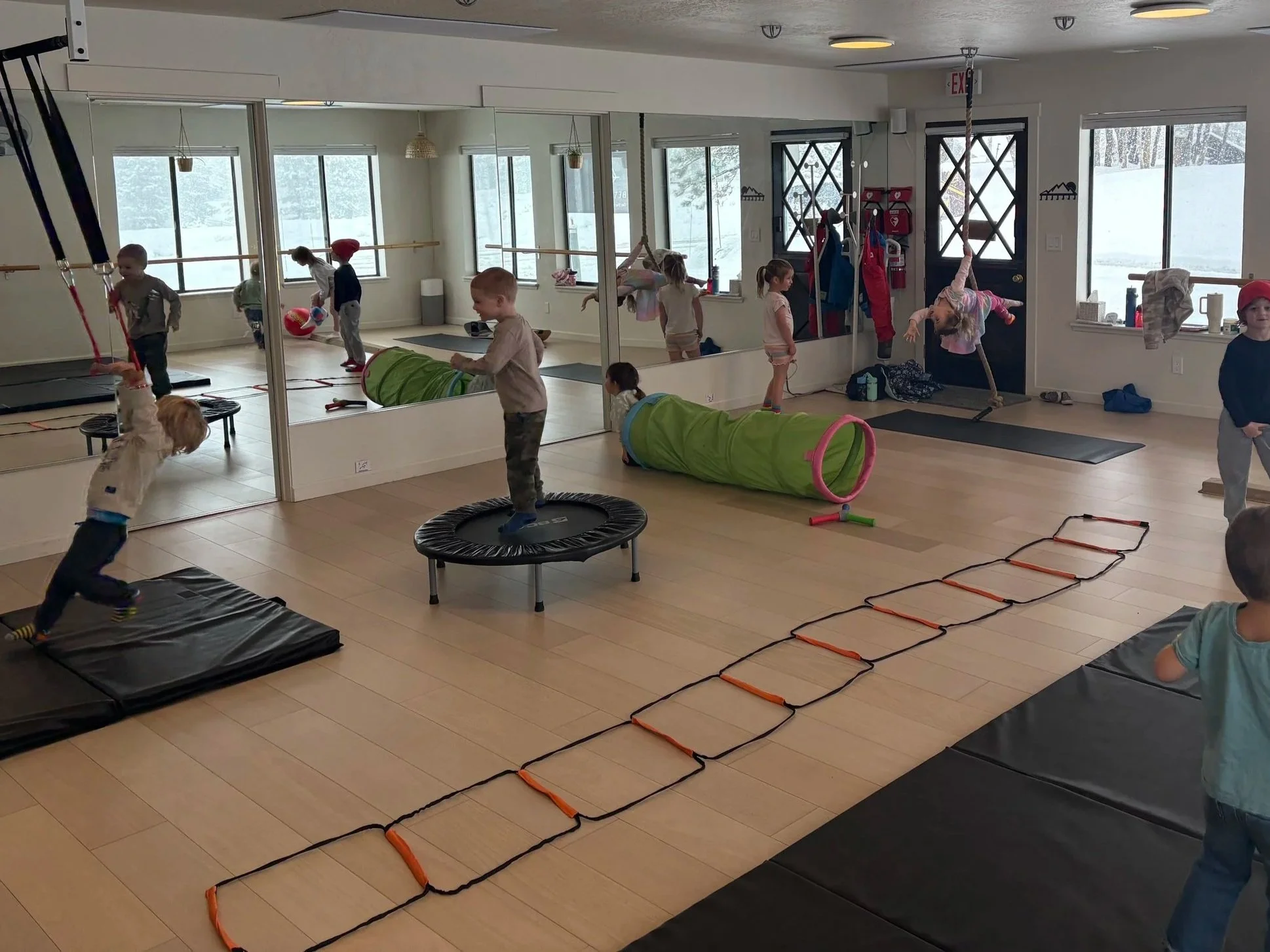 Children playing in an indoor playground, engaging in activities such as jumping on trampolines, crawling through tunnels, and climbing agility ladders, with large windows showing a snowy landscape outside.