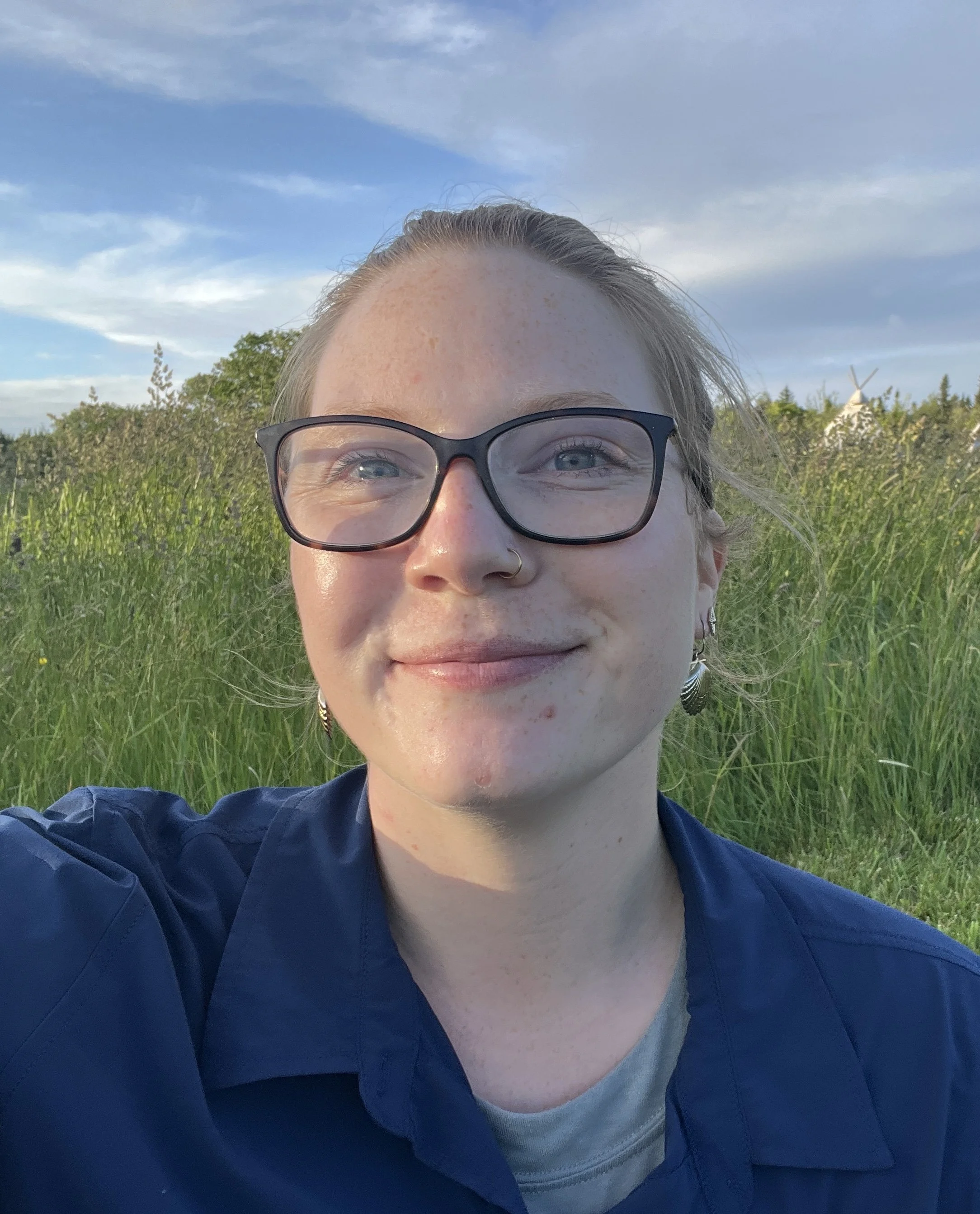young blonde woman sits in a field of grass and smiles at the camera