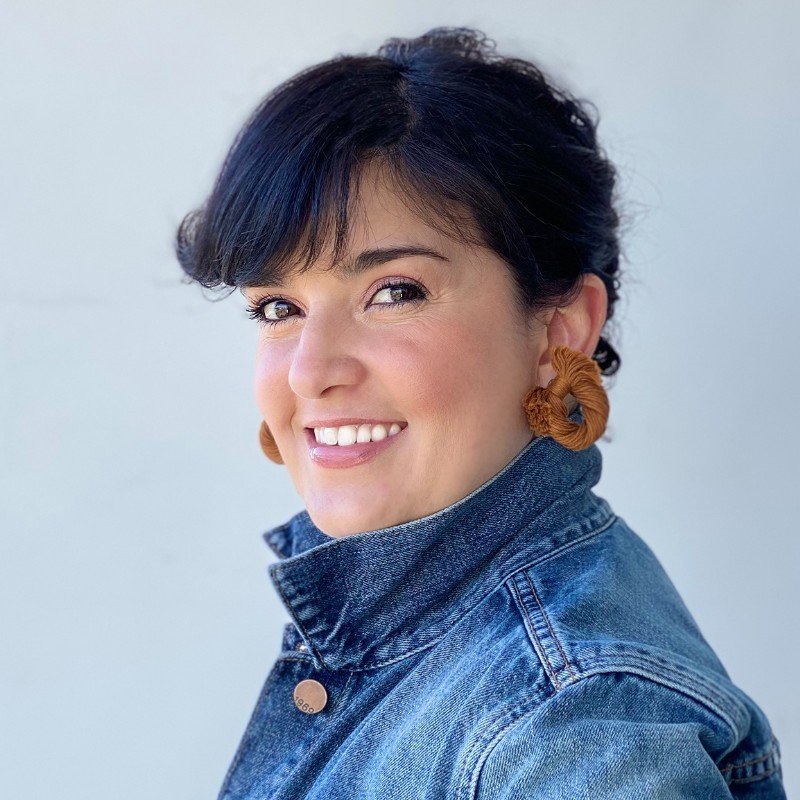 Person smiling, wearing a denim jacket and beige hoop earrings.