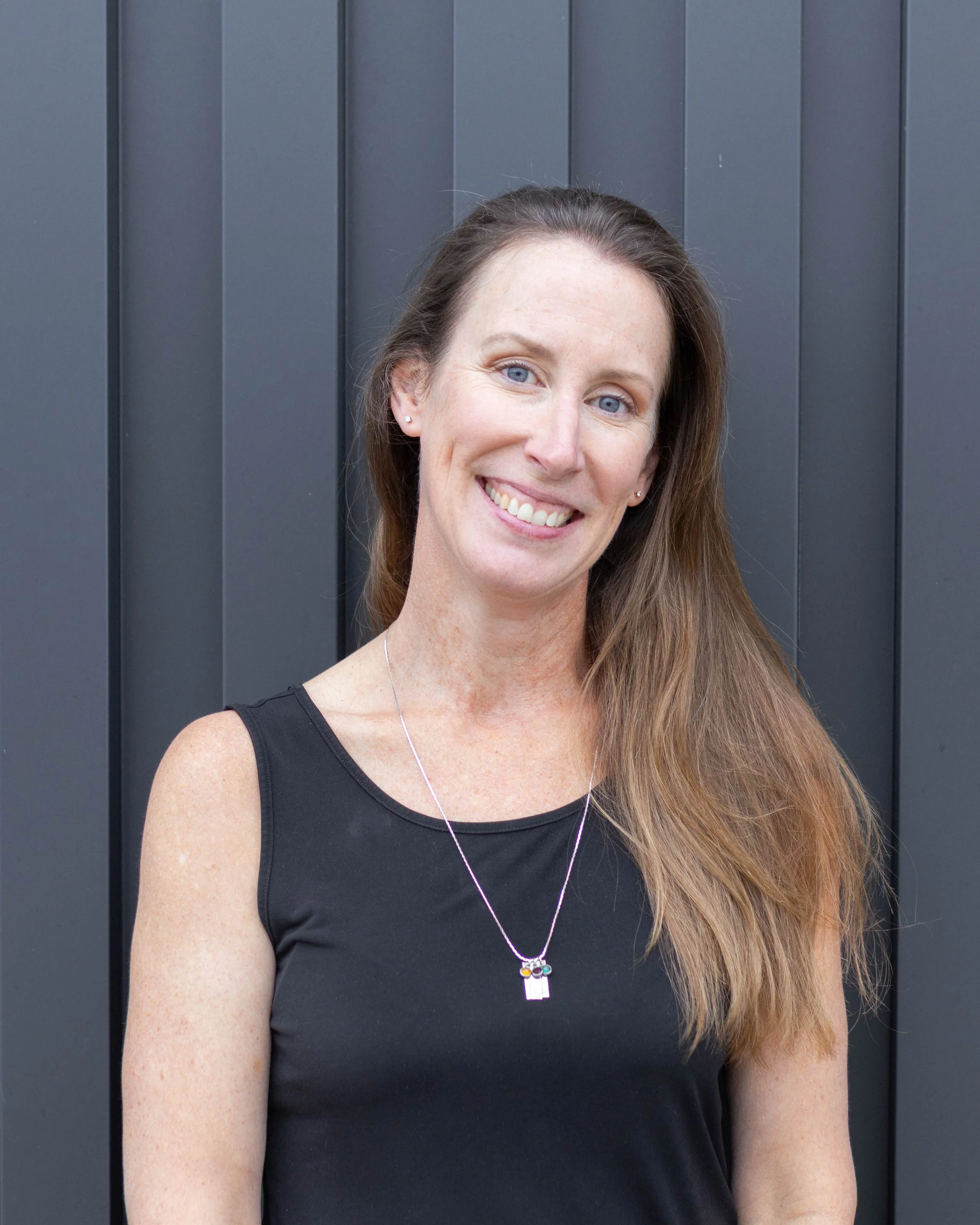 A smiling woman with long brown hair Wearing a black sleeveless top and a necklace with a small pendant standing in front of a dark grey wooden fence.