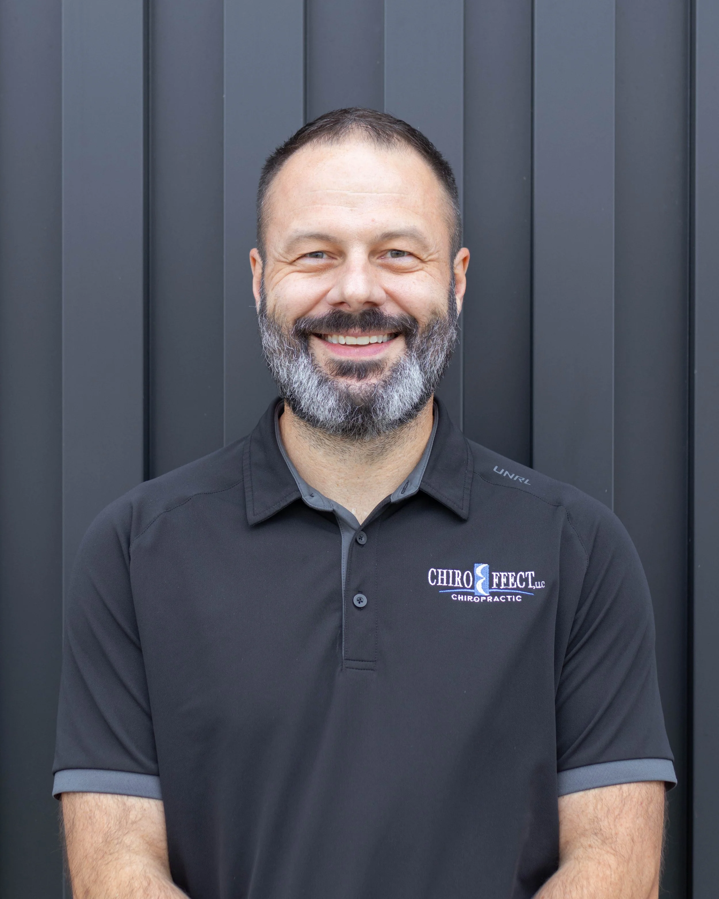 A man with a beard smiling, wearing a black polo shirt with logo, standing in front of a dark gray wall.