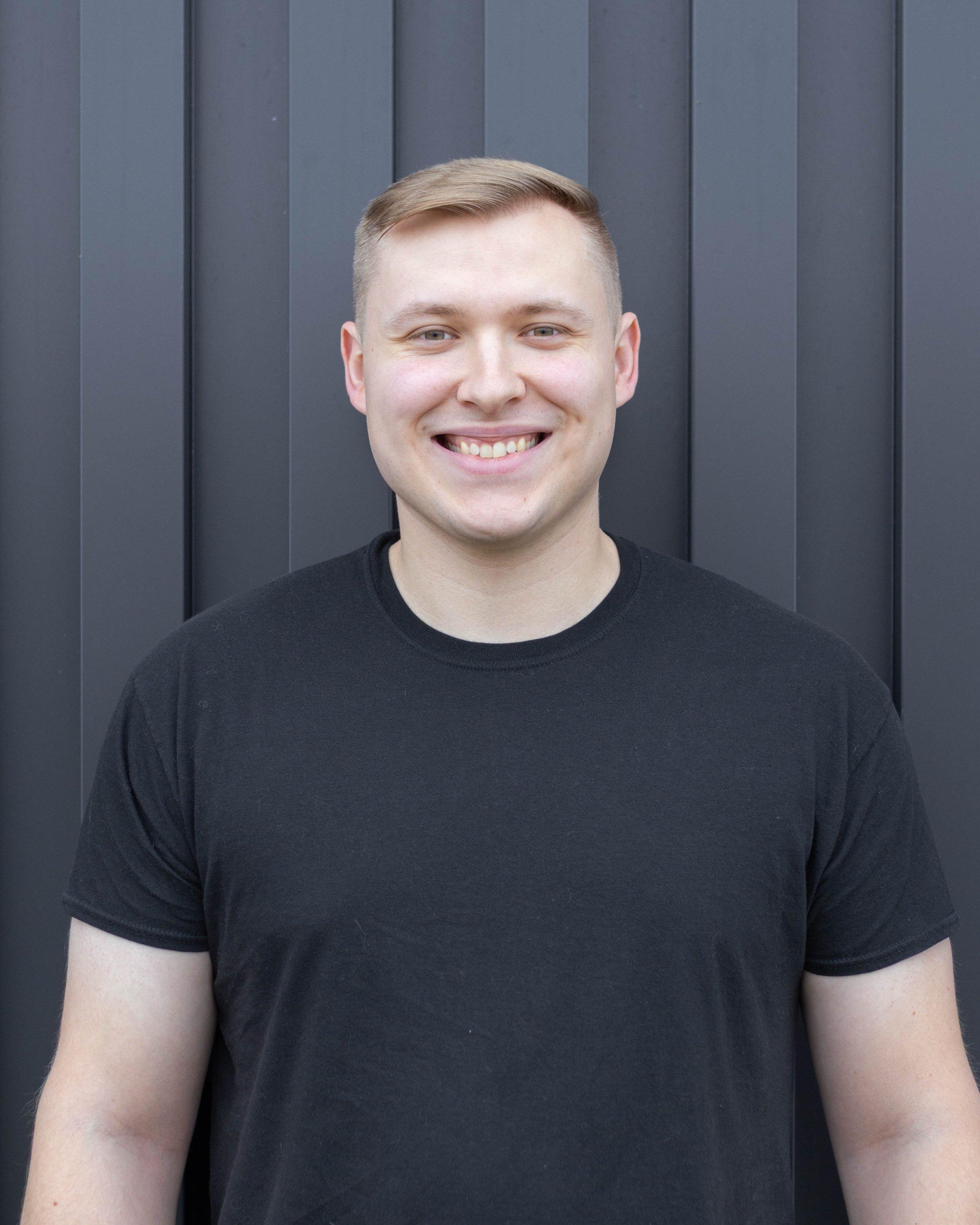 A young man with blonde hair smiling and wearing a black t-shirt, standing in front of a dark gray wooden wall.