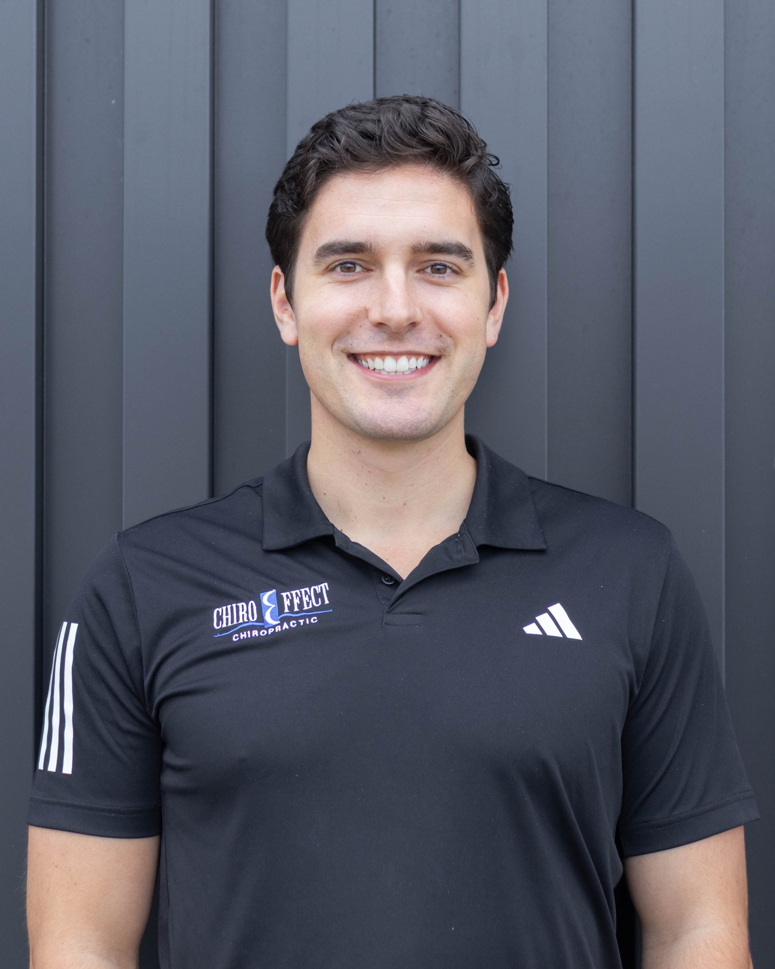 A young man with dark hair and a bright smile wearing a black polo shirt with chiropractic branding, standing in front of a dark gray wall.