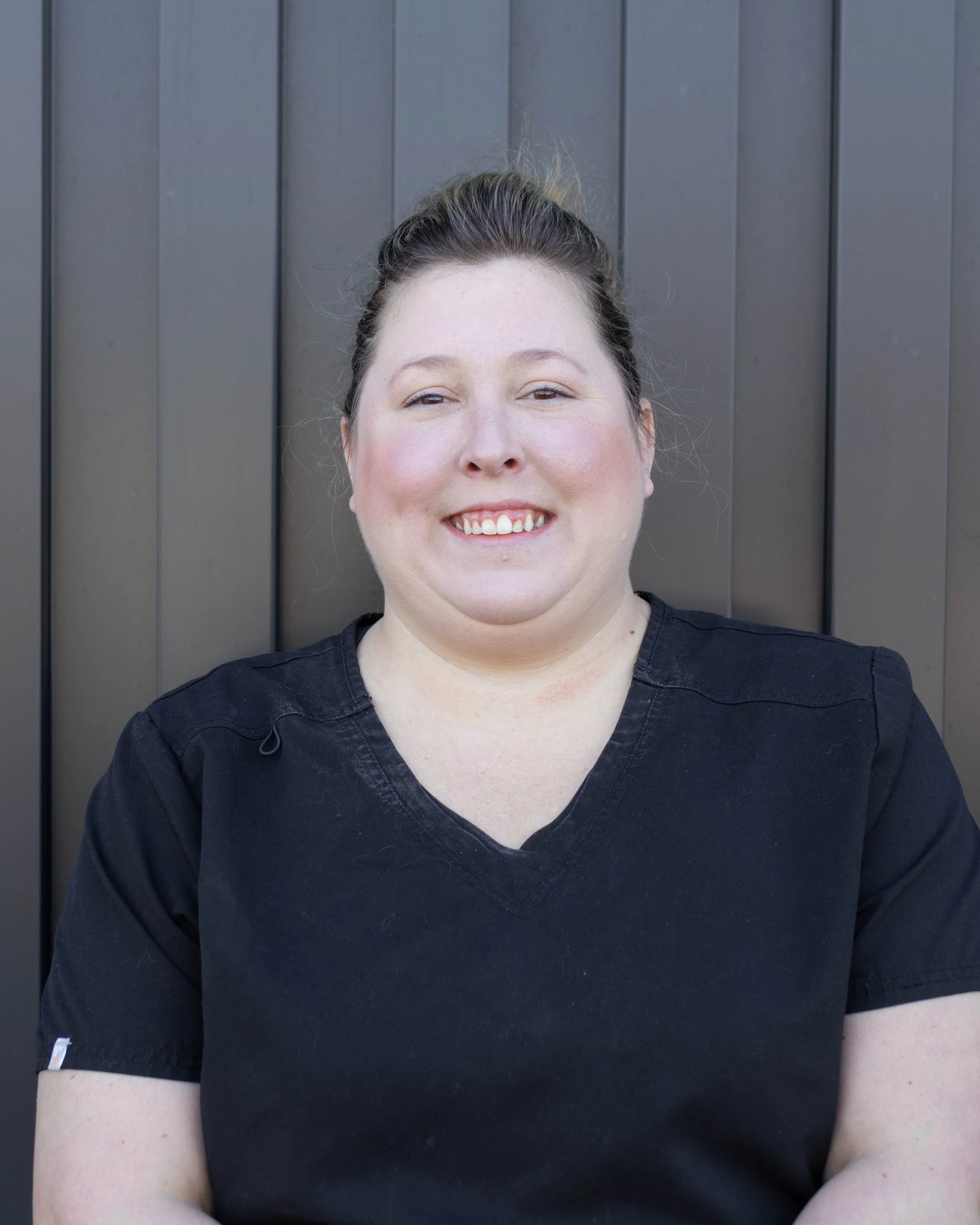 A woman with dark hair in a bun, smiling, wearing a black top, standing against a dark-colored vertical panel background.