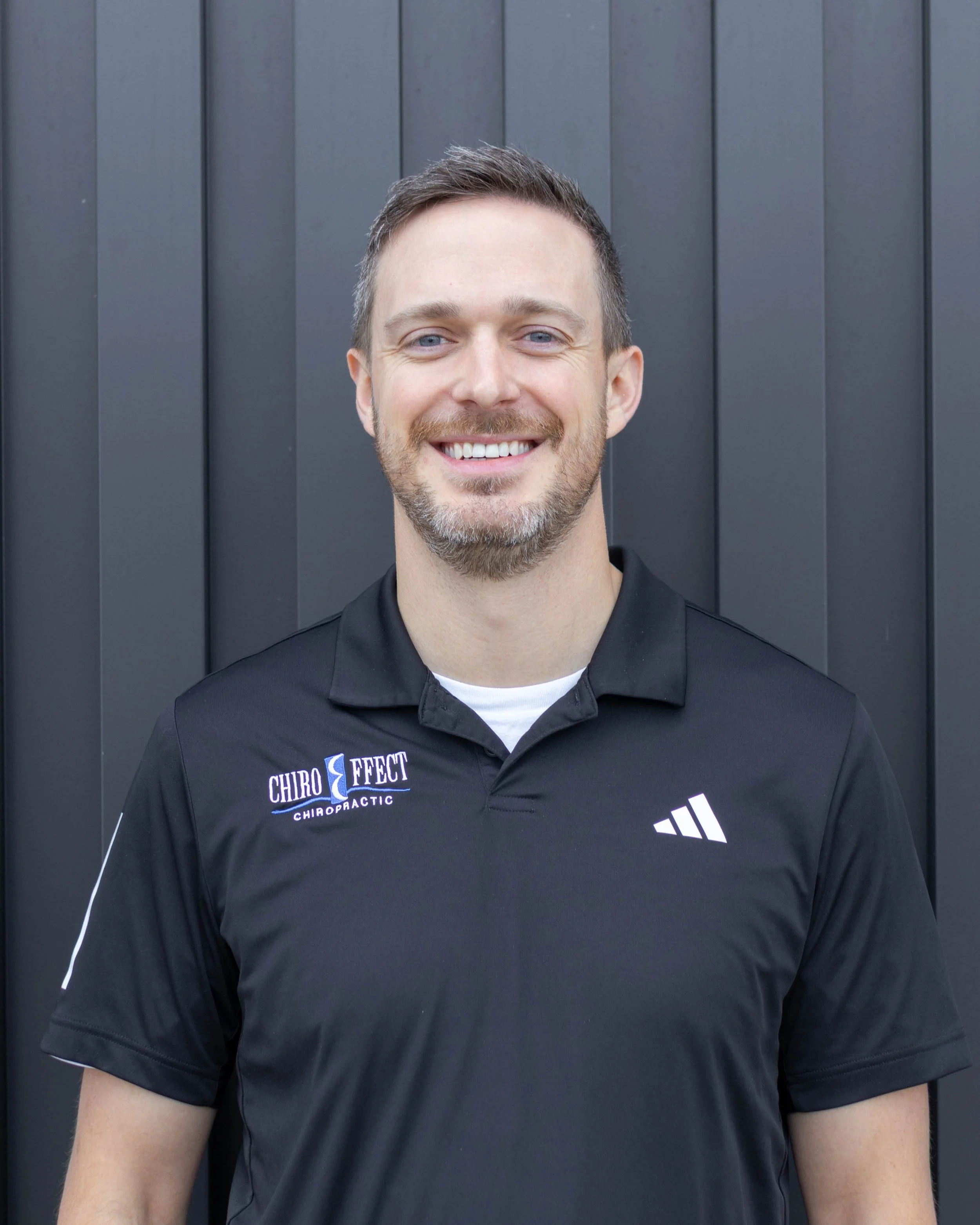 A smiling man in a black polo shirt with chiropractor branding, standing outdoors in front of a dark gray wooden wall.