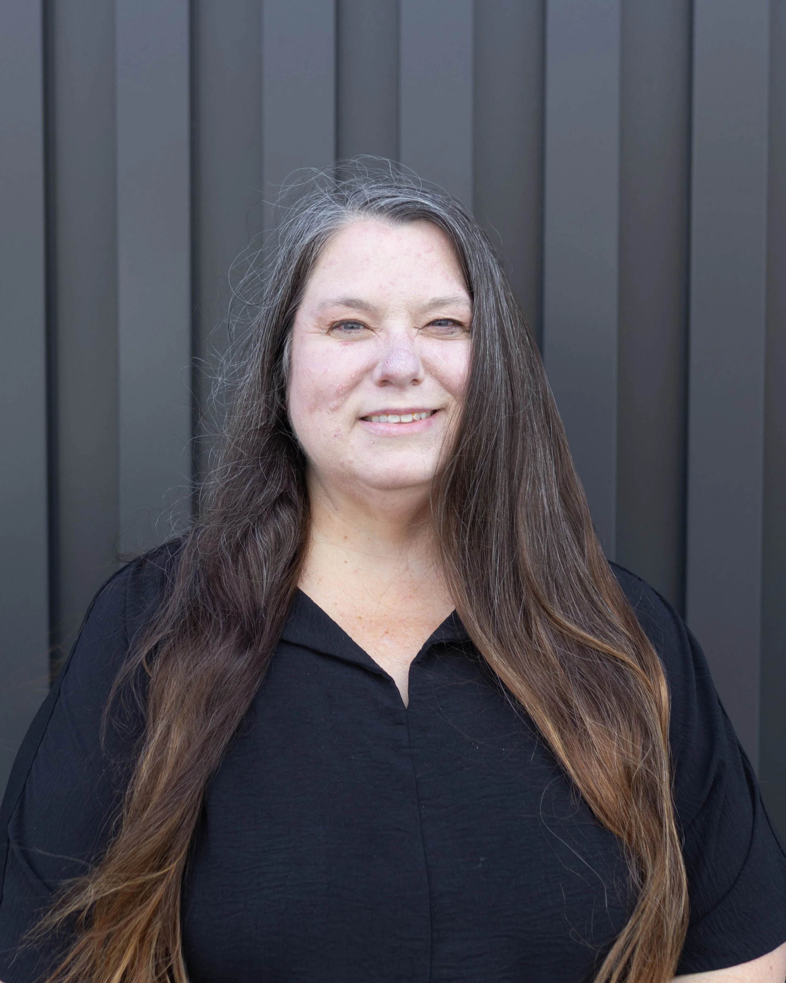 Portrait of a woman with long brown hair standing outdoors in front of a dark gray wooden fence, smiling slightly, wearing a black shirt.