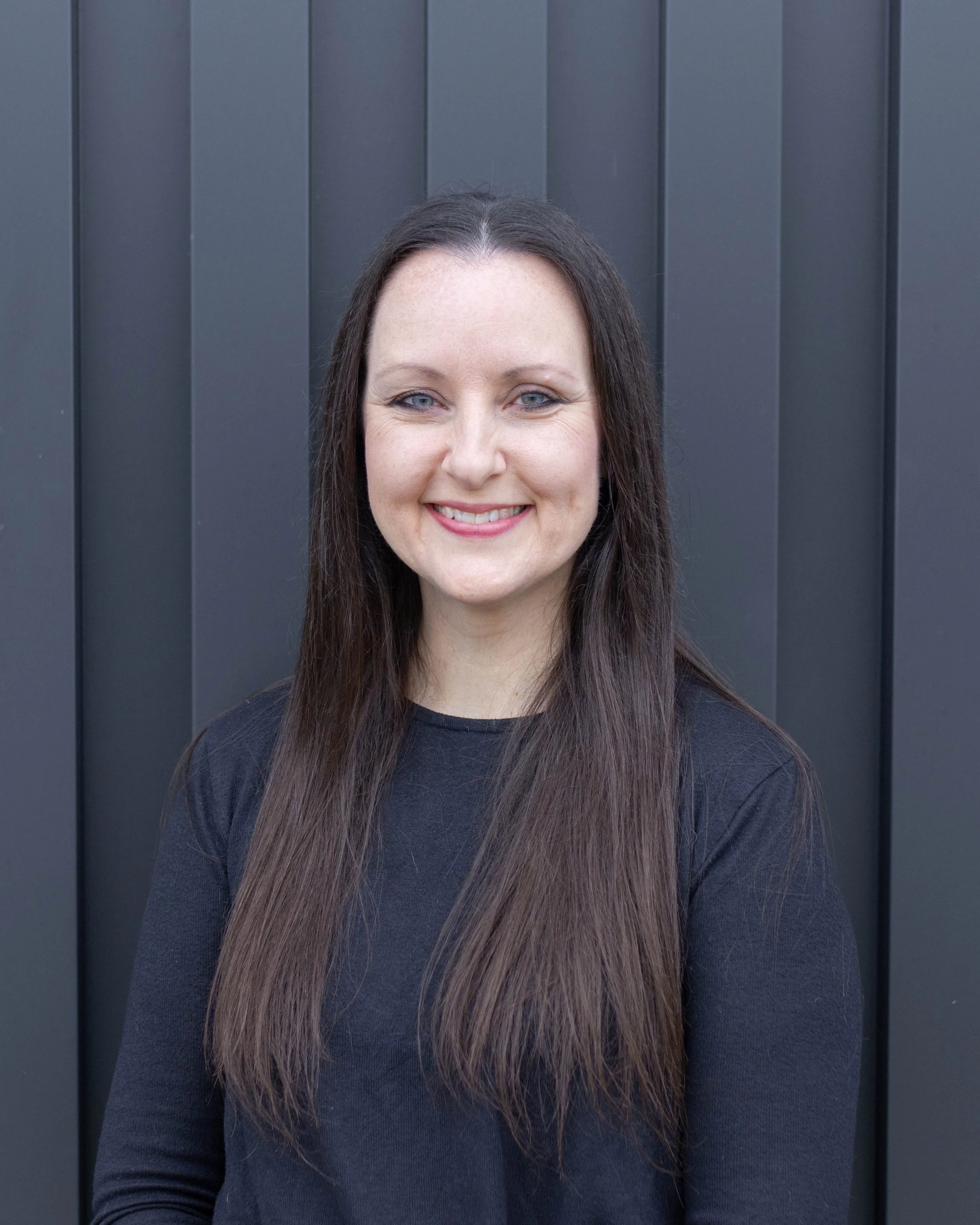 Portrait of a woman with long dark hair, wearing a black top, smiling, standing in front of a dark gray wall.