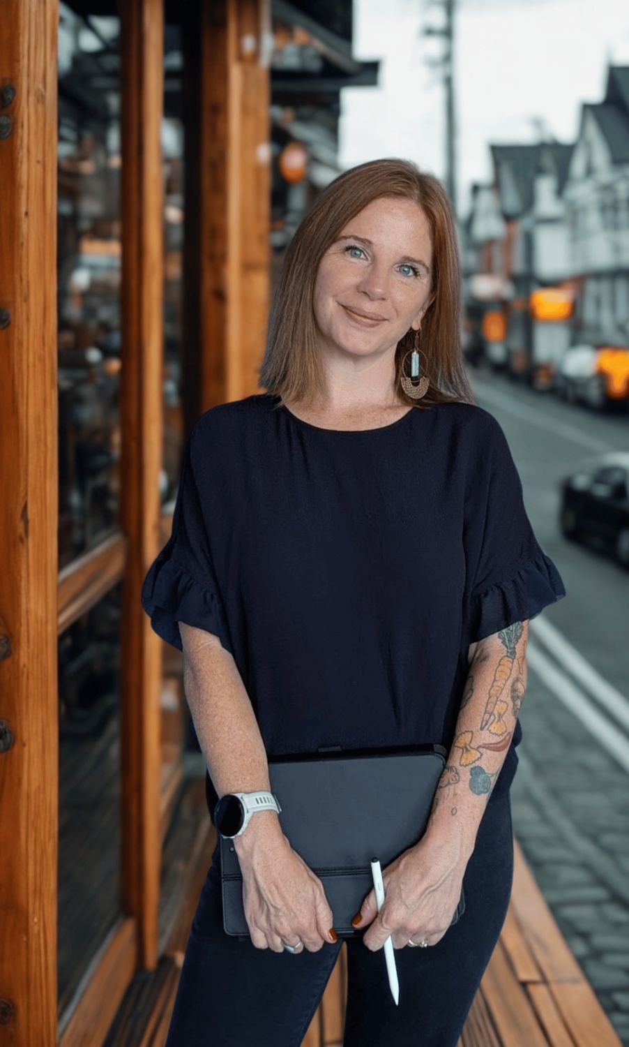 A woman with red hair and blue eyes smiling, standing outside near a wooden structure on a street with parked cars and buildings in the background. She is wearing a black dress with ruffled sleeves, earrings, a white watch, and holding a black tablet and stylus.