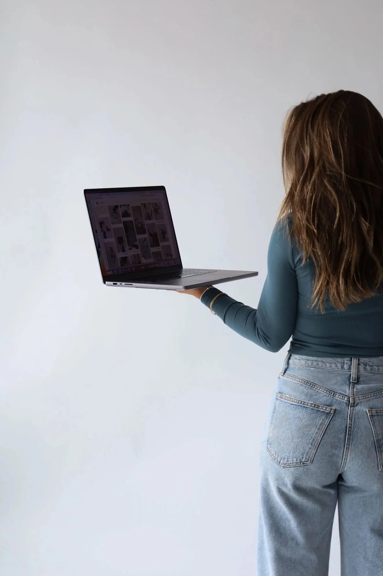 A woman with long wavy hair in a teal long-sleeve shirt and light blue jeans is holding a laptop in her right hand against a plain white background.