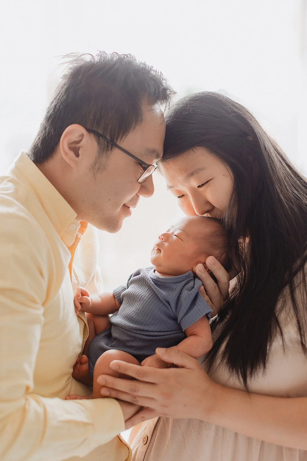 Mother and Father stand by a window holding their newborn baby for their newborn baby shoot with Kith and Kin photography.