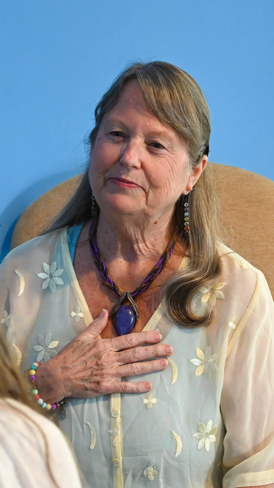An elderly woman sitting on a chair with a blue background, wearing a floral sheer blouse, colorful jewelry, and a purple gemstone necklace, with her hand on her chest.