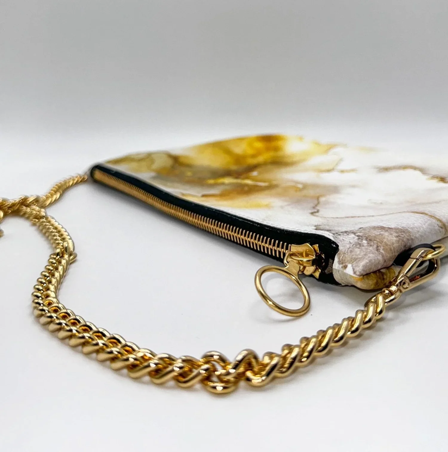 Close up photo of a white and gold patterned clutch bag with a gold chain strap, lying on a white-grey surface.