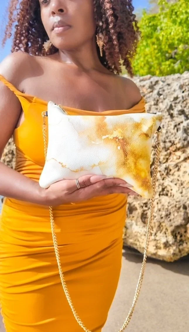 A model holding a white and gold patterned clutch bag with a gold chain strap. The model is wearing a gold dress, she has curly hair and is standing outside on a beach.