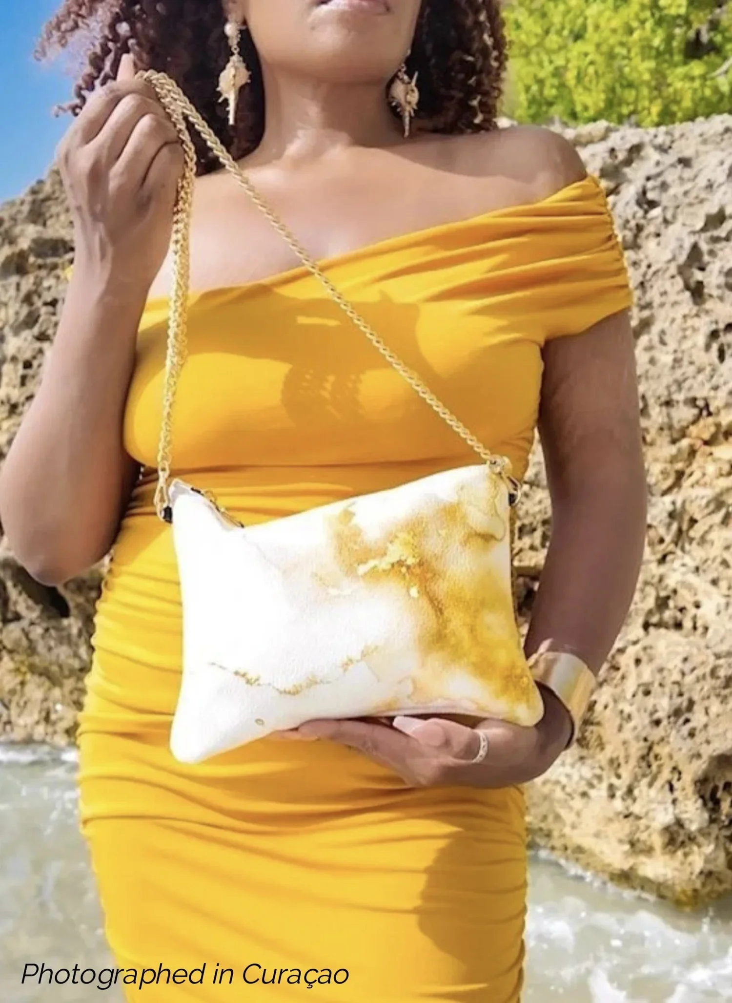 A model holding a white and gold patterned clutch bag with a gold chain strap. The model is wearing a gold dress, she has curly hair and is standing outside on a beach.