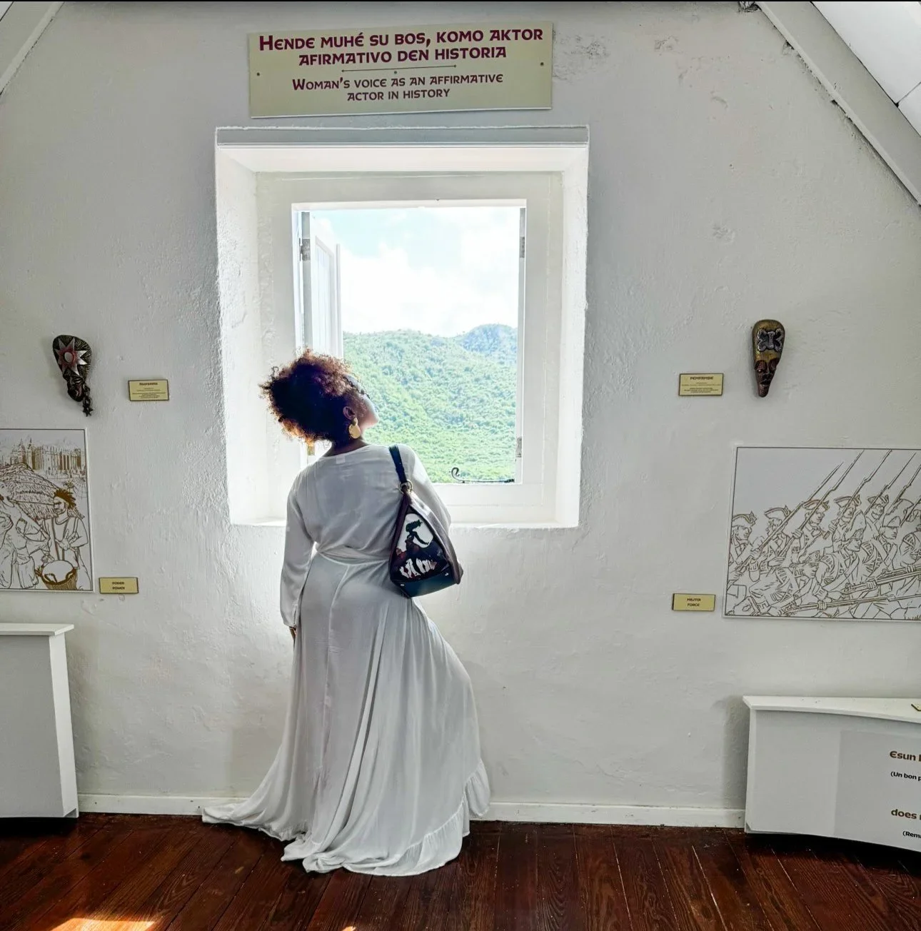 A model wearing a white dress holding a maroon-colored leather shoulder bag with a unique graphic print of a moon goddess connected with her cycle. The sign behind her in Dutch translates to "WOMAN'S VOICE AS AN AFFIRMATIVE ACTOR IN HISTORY."