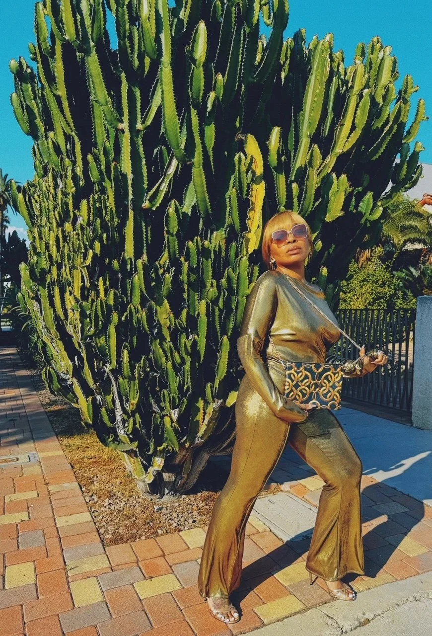 A model wearing a shiny gold top and pants, holds a black and gold print leather purse with a long gold chain, while standing outside with a large cacti green tree behind her.