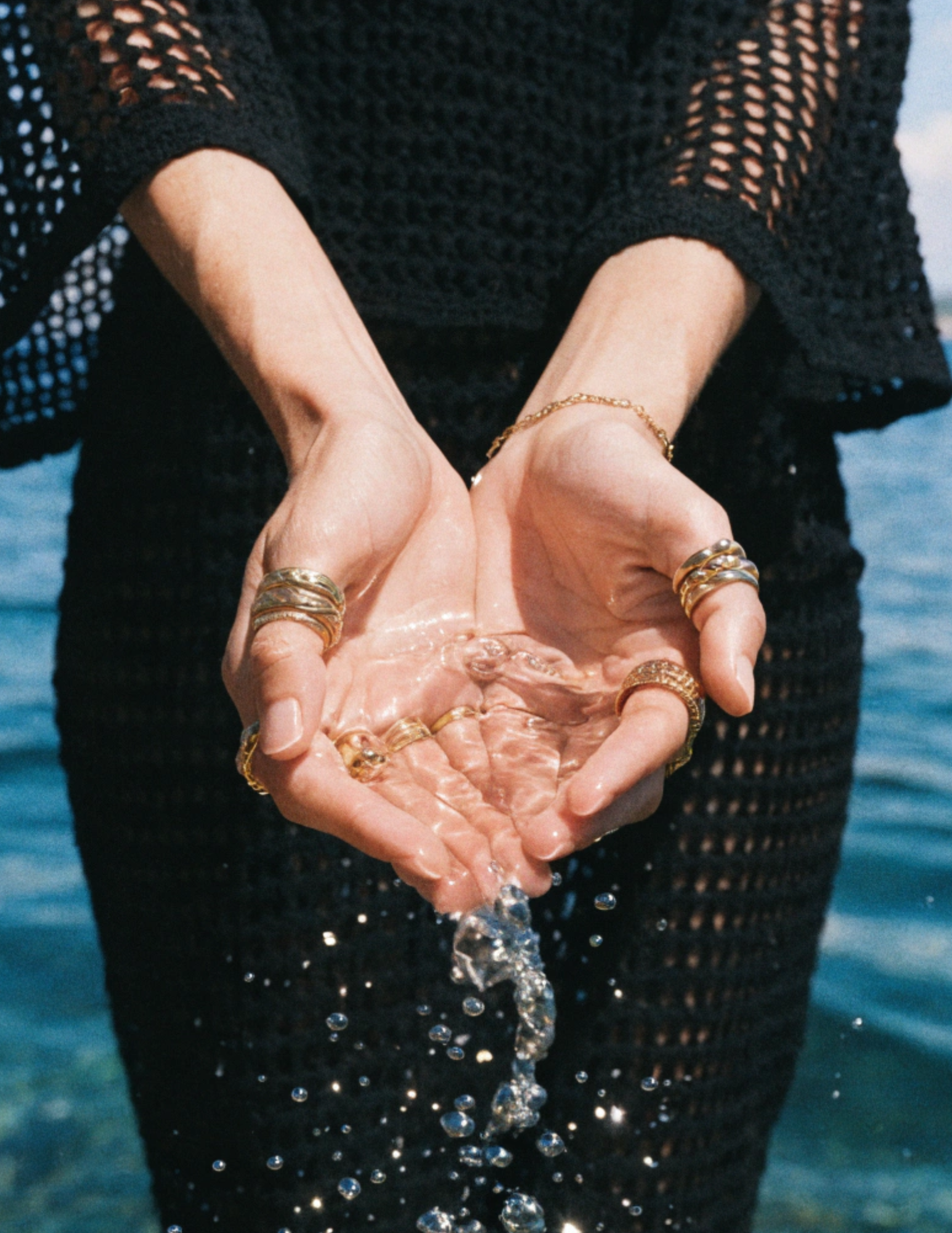 Close up photo of a woman standing on a beach, the ocean behind her, the sun shines brightly. Her hands are cupped together holding ocean water that spills in slow motion from her hands. She  wears a black crochet dress.