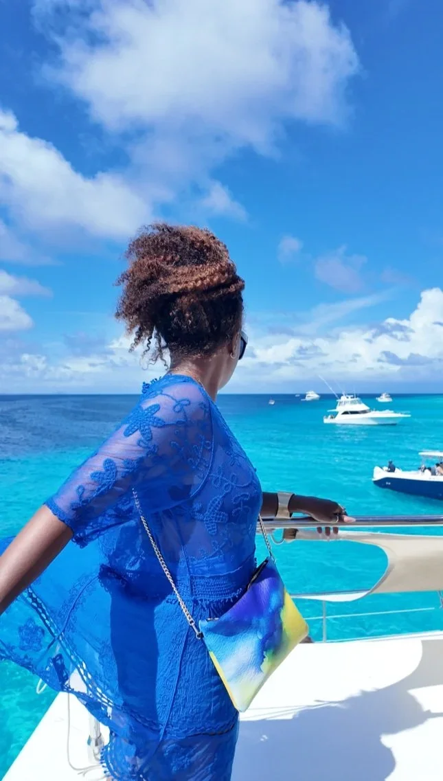 A model with curly hair wearing a blue dress, standing on a yacht looking at the blue ocean behind her, wearing a yellow and blue crossbody purse, with a gold chain.