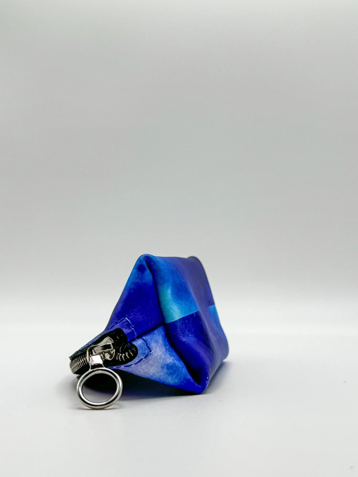 A close up photo of the side and bottom of a blue-purple multicolored leather shell coin purse, with a silver zipper and circle puller, on a white background.