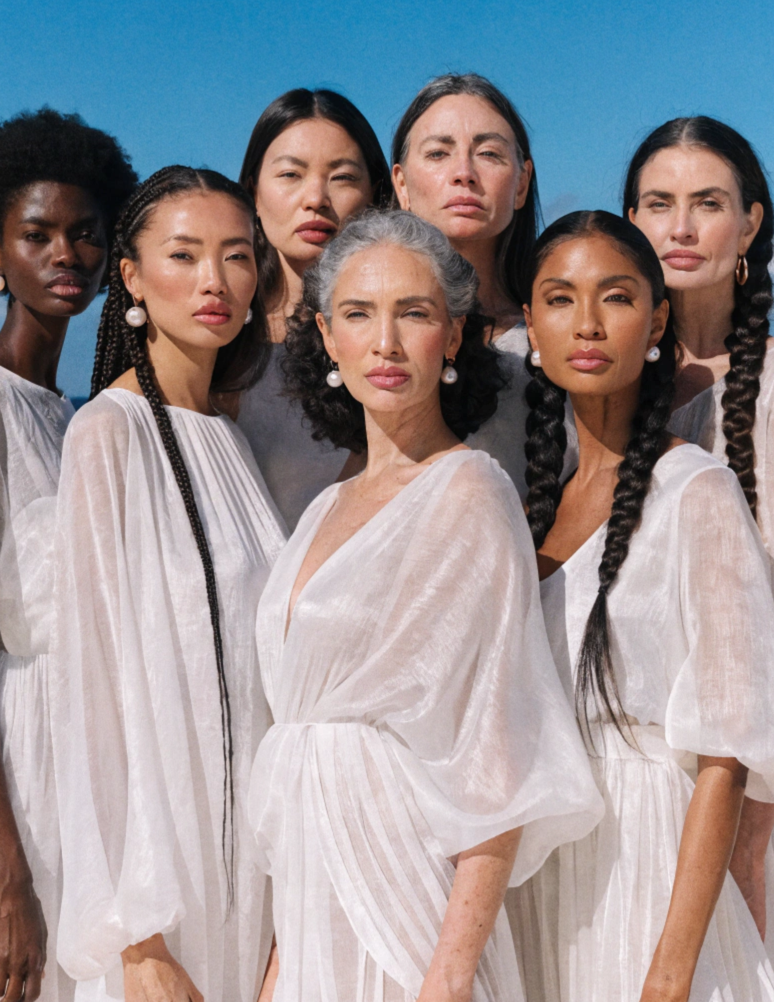A diverse group of women stand together dressed in white dresses. They all range in heights, age, ethnicity, and skin tones.