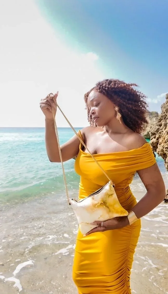 A model holding a white and gold patterned clutch bag with a gold chain strap. The model is wearing a gold dress, she has curly hair and is standing outside on a beach.