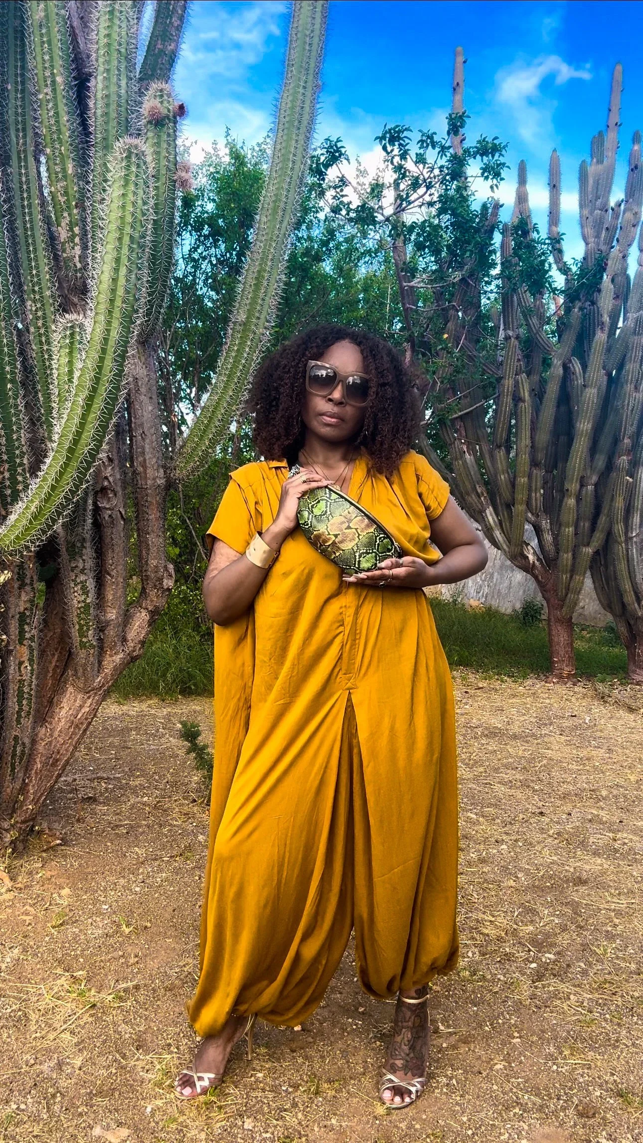 A photo of a model with curly hair wearing a yellow jumpsuit, and a green and black snakeskin print fanny pack across her body. The model is standing outside surrounded by green cacti and a blue sky with clouds.