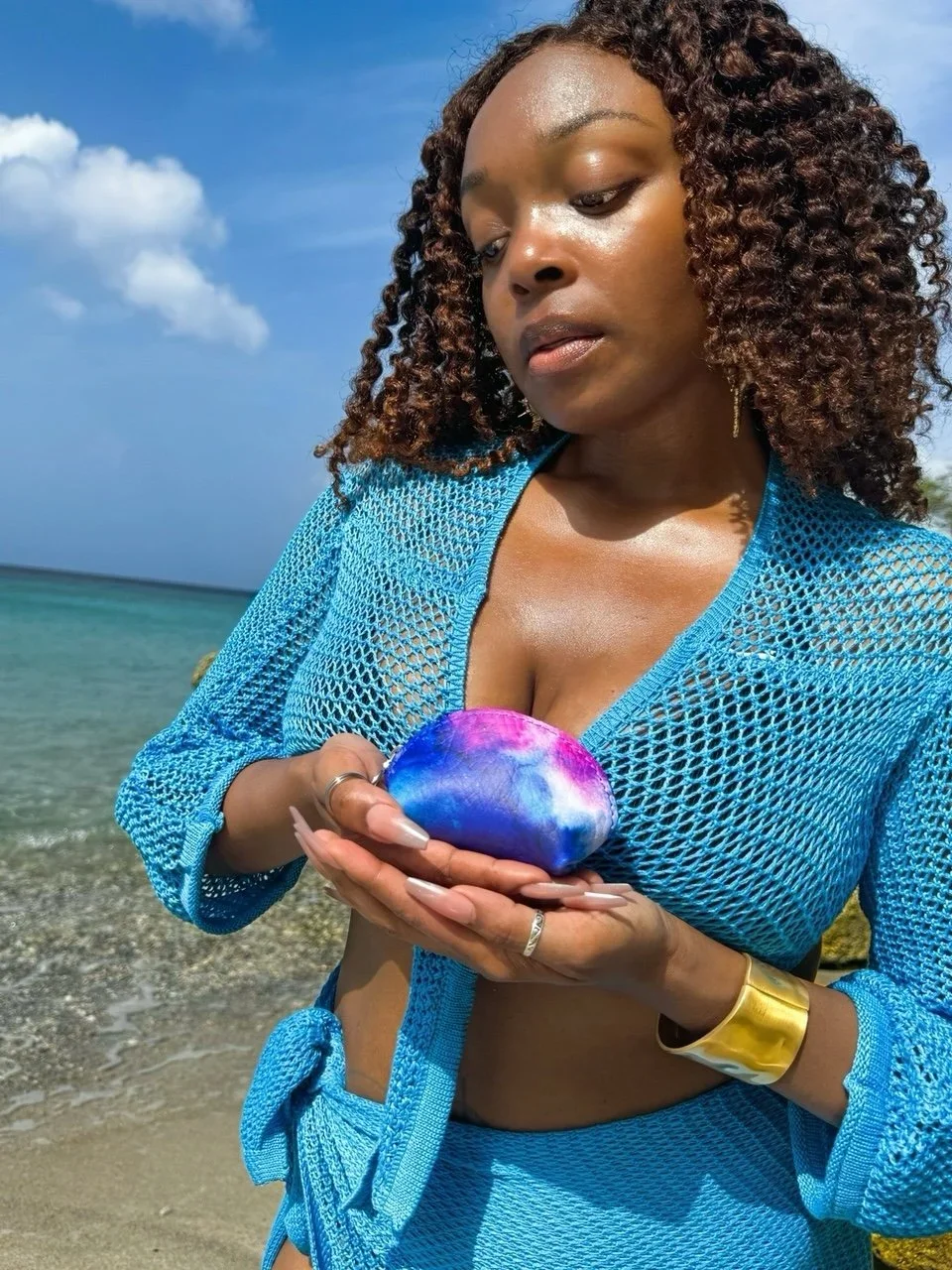 A model standing on a beach holding a blue-purple multicolored leather shell coin purse, wearing a blue crochet knit two-piece set.