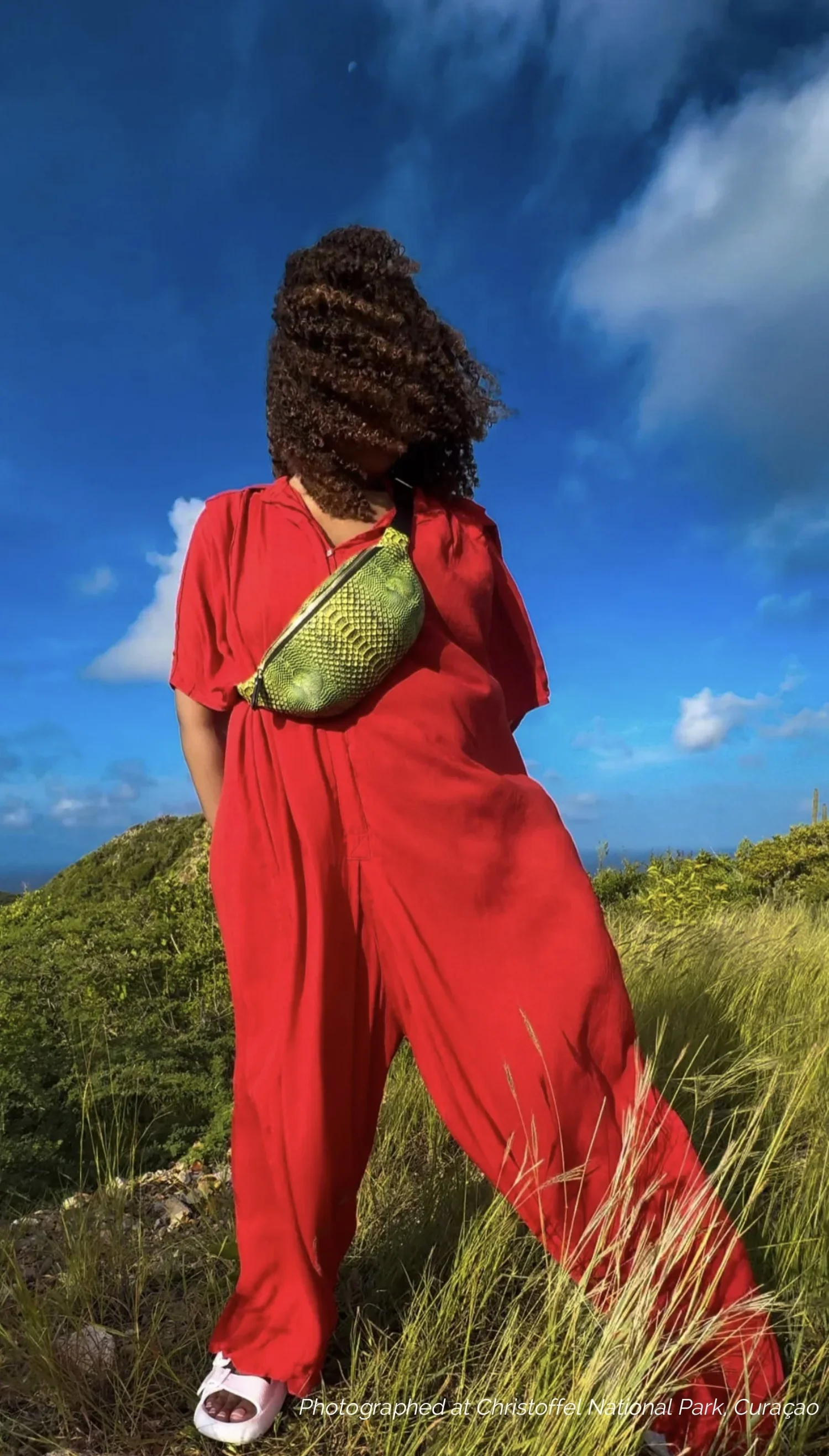 A model with curly hair, wearing a red jumpsuit with a bright green Fanny Pack across her body, against a bright blue sky with white clouds..