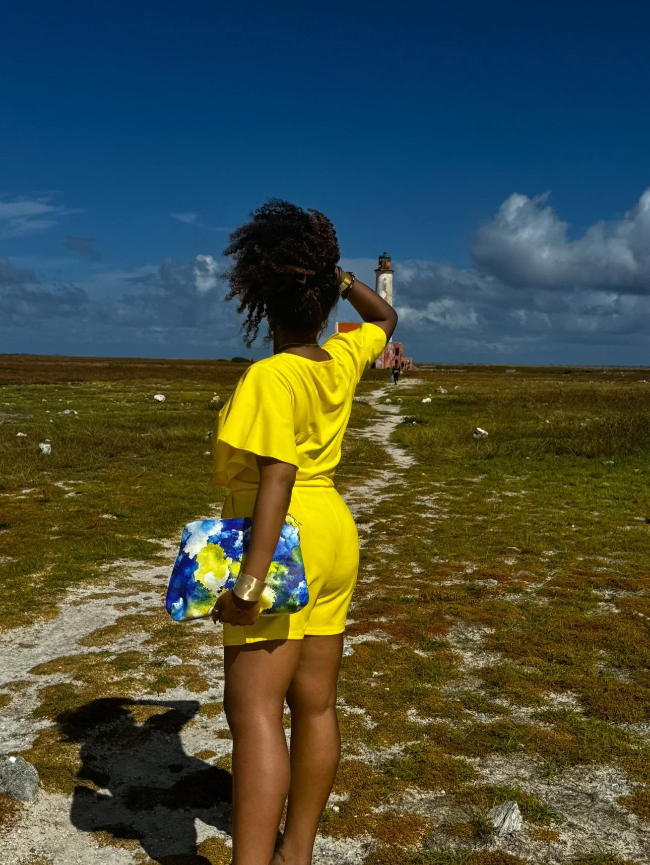 A model wearing a yellow shirt and shirt holding a large yellow, blue and white leather clutch in front of the iconic pink lighthouse on Klein Curaçao.