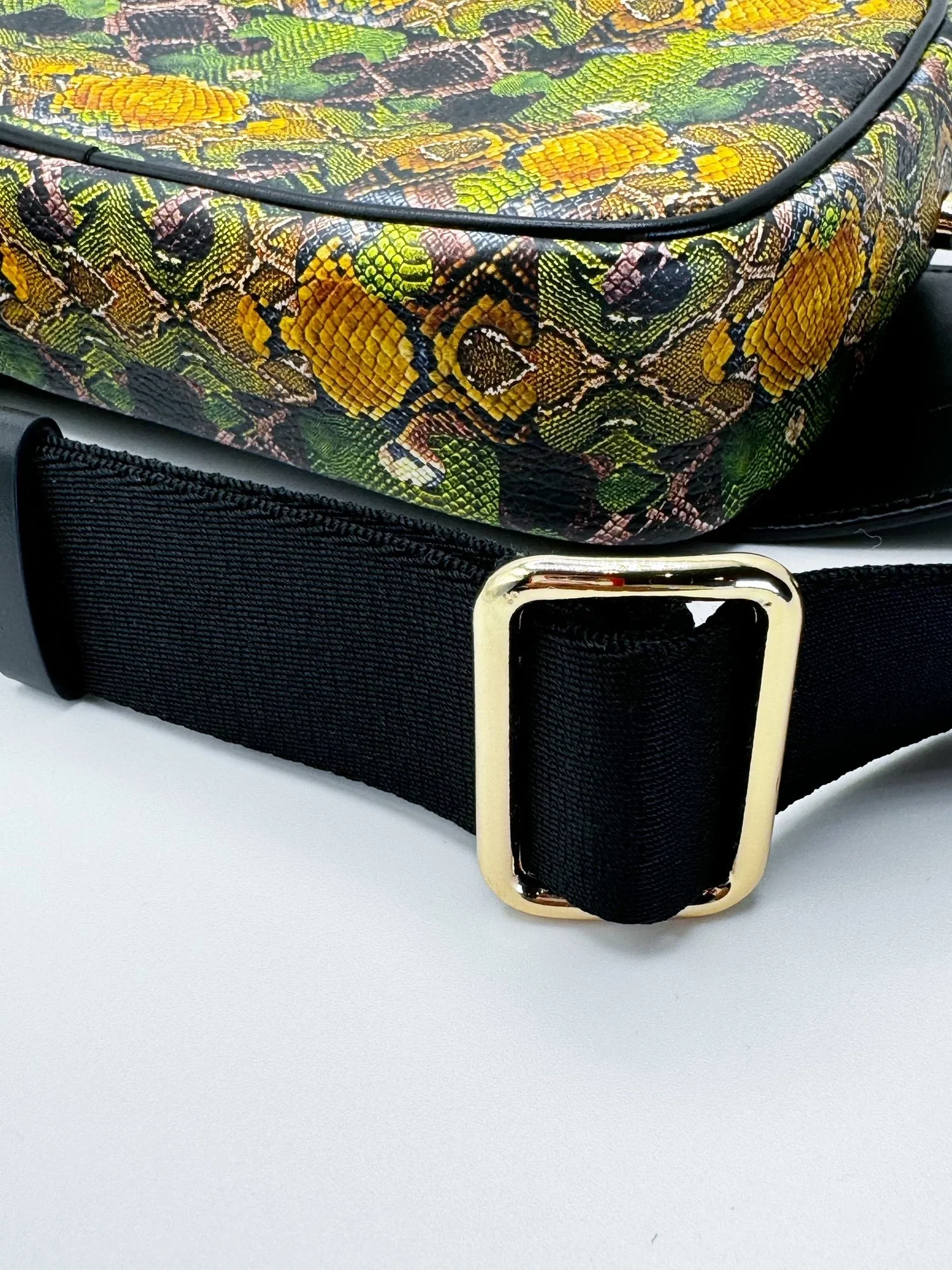 A close up photo of a multicolored Leather bag with a black waist strap, a black leather zipper pull and gold zipper. The background is white-grey.