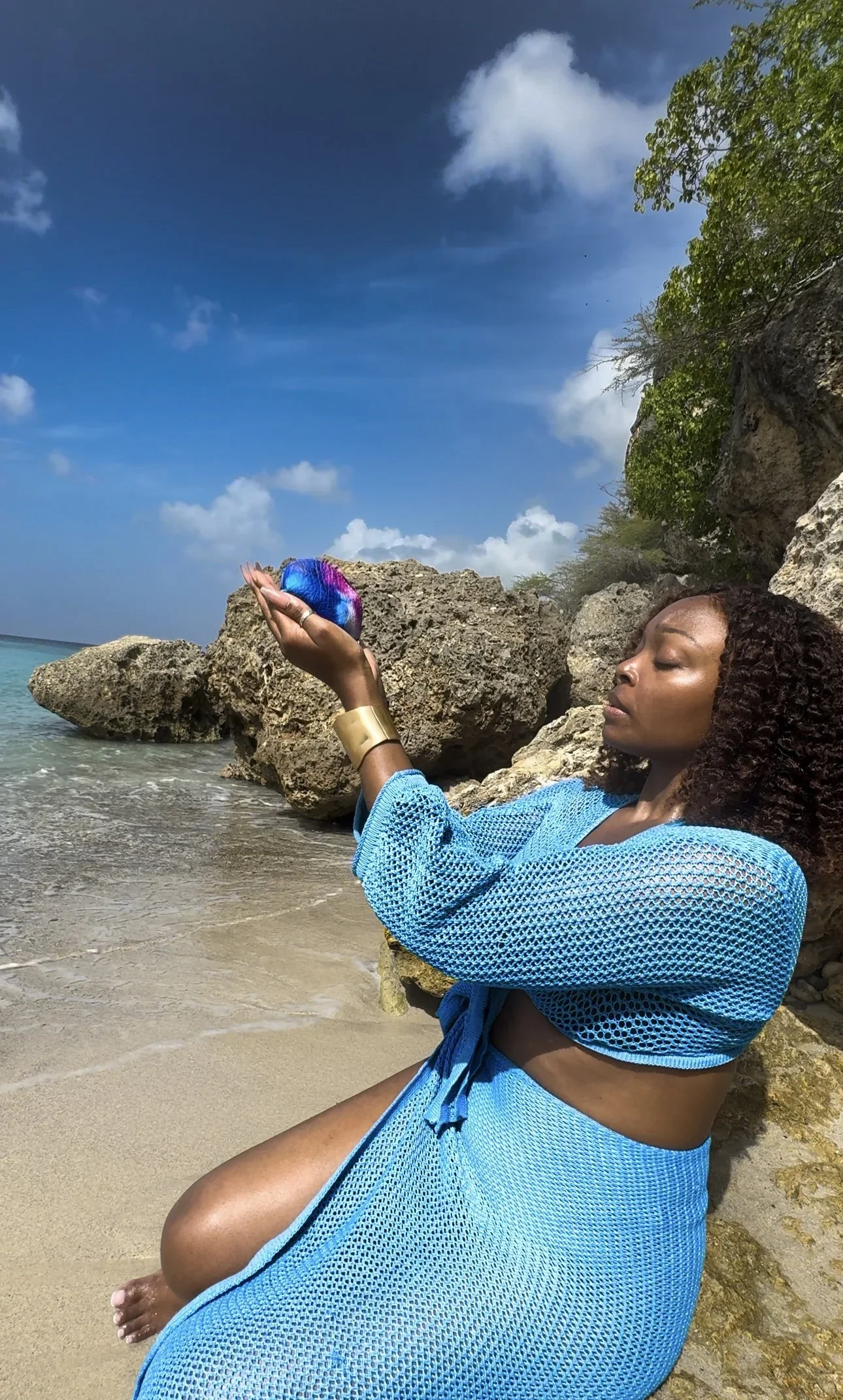 A model standing on a beach holding a blue-purple multicolored leather shell coin purse, wearing a blue crochet knit two-piece set.