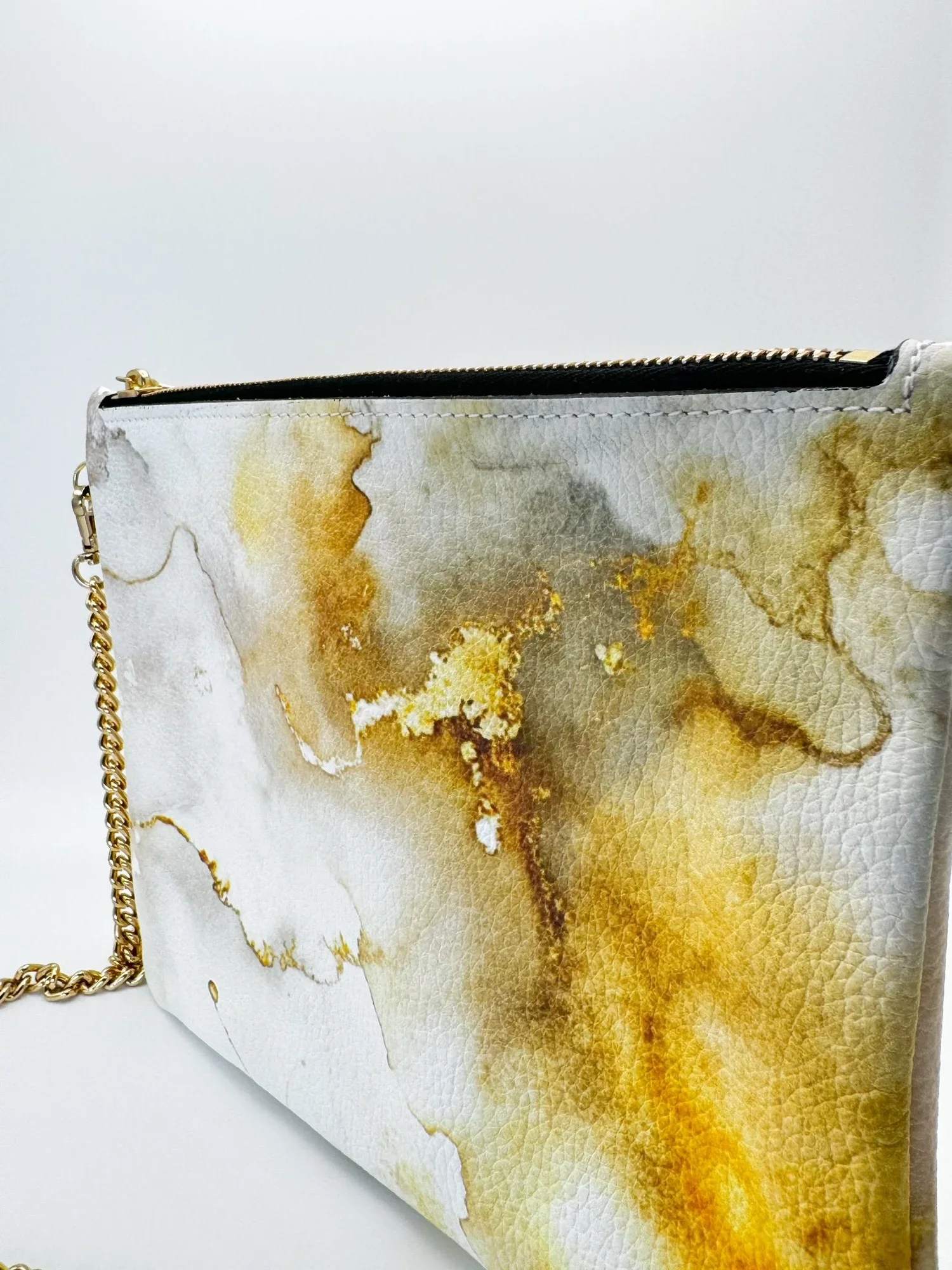 Close up photo of a white and gold patterned clutch bag with a gold chain strap, lying on a white-grey surface.