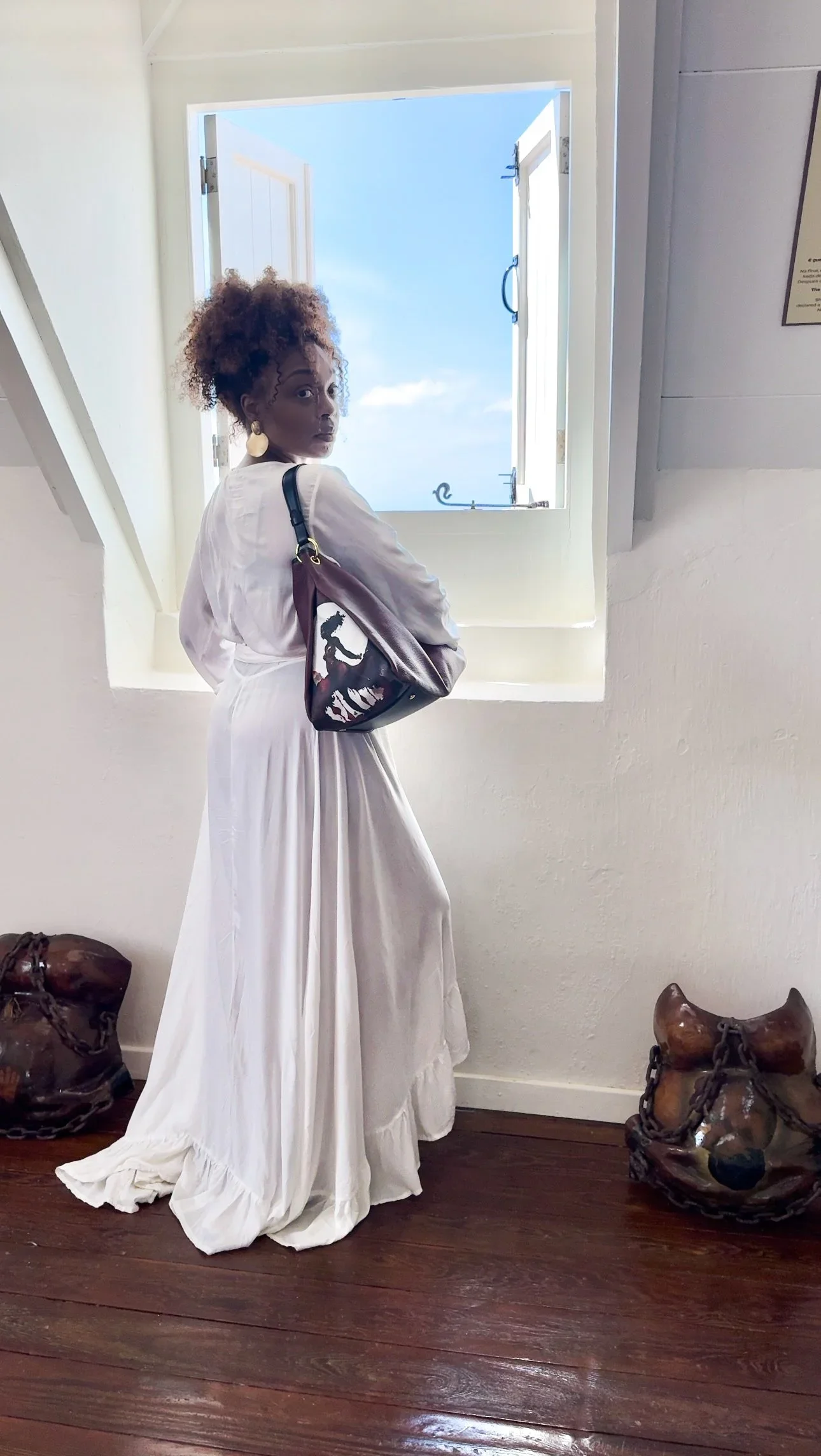 A model wearing a white dress holding a maroon-colored leather shoulder bag with a unique graphic print of a moon goddess connected with her cycle.