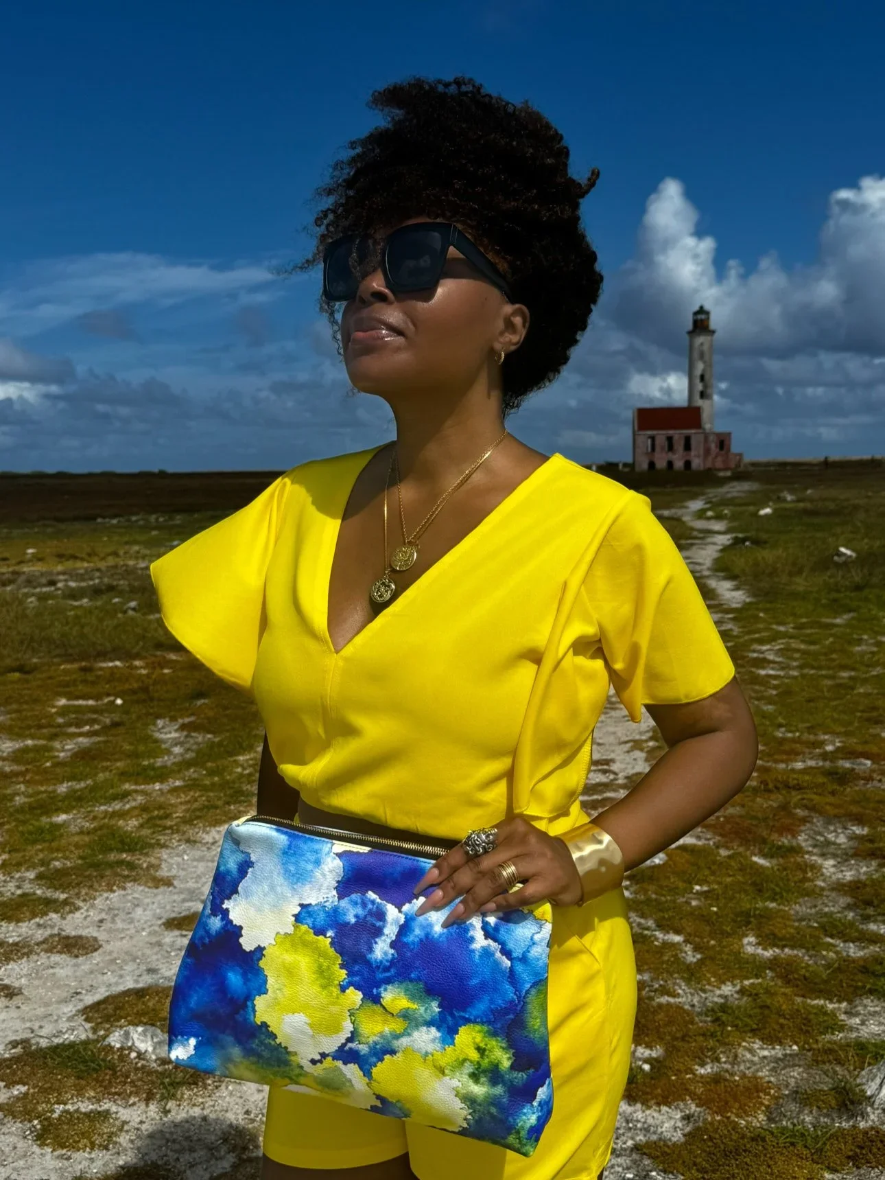 A model wearing a yellow shirt and shirt holding a large yellow, blue and white leather clutch in front of the iconic pink lighthouse on Klein Curaçao.