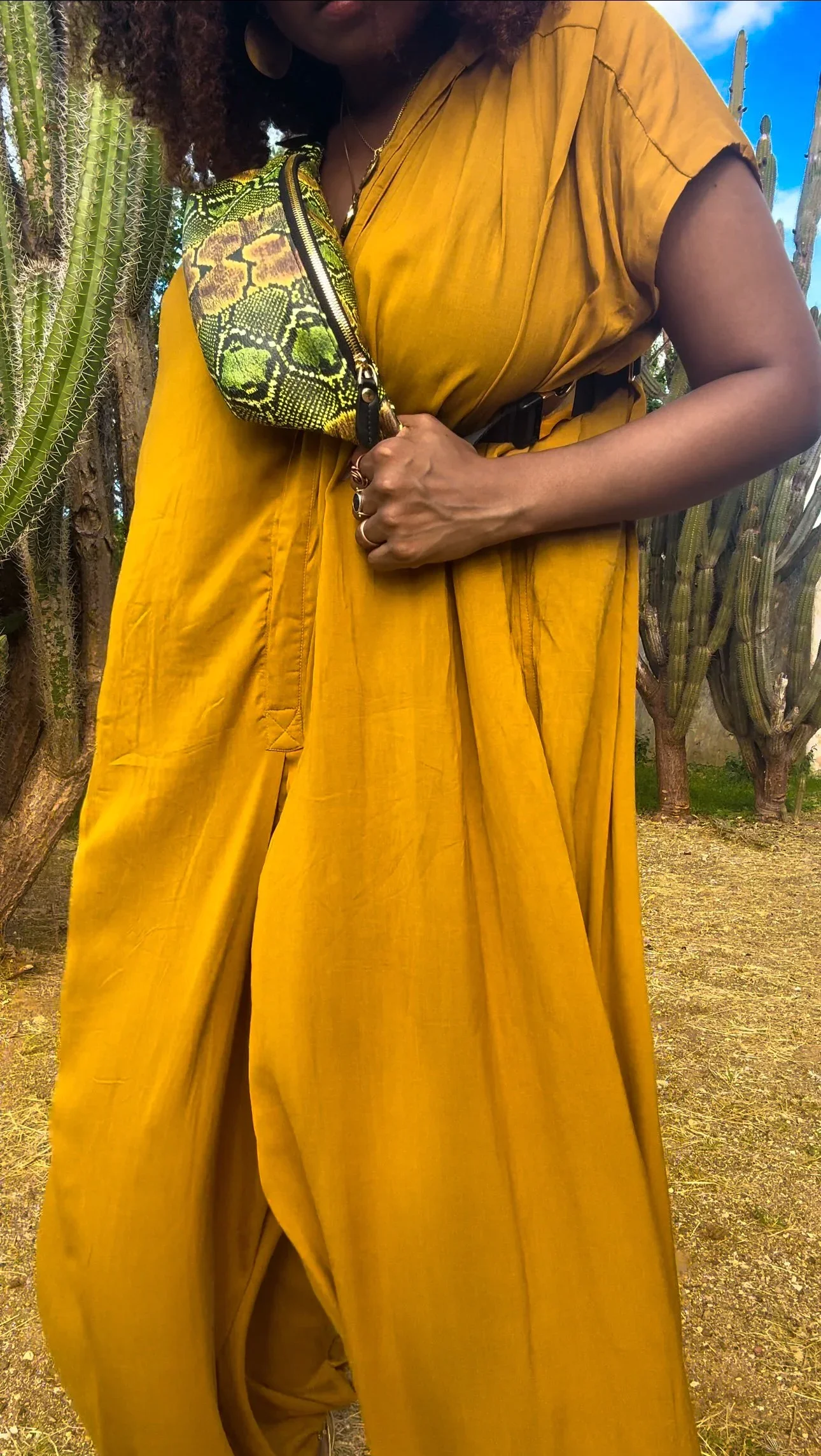 A photo of a model wearing a yellow jumpsuit, and a green and black snakeskin print fanny pack across her body.