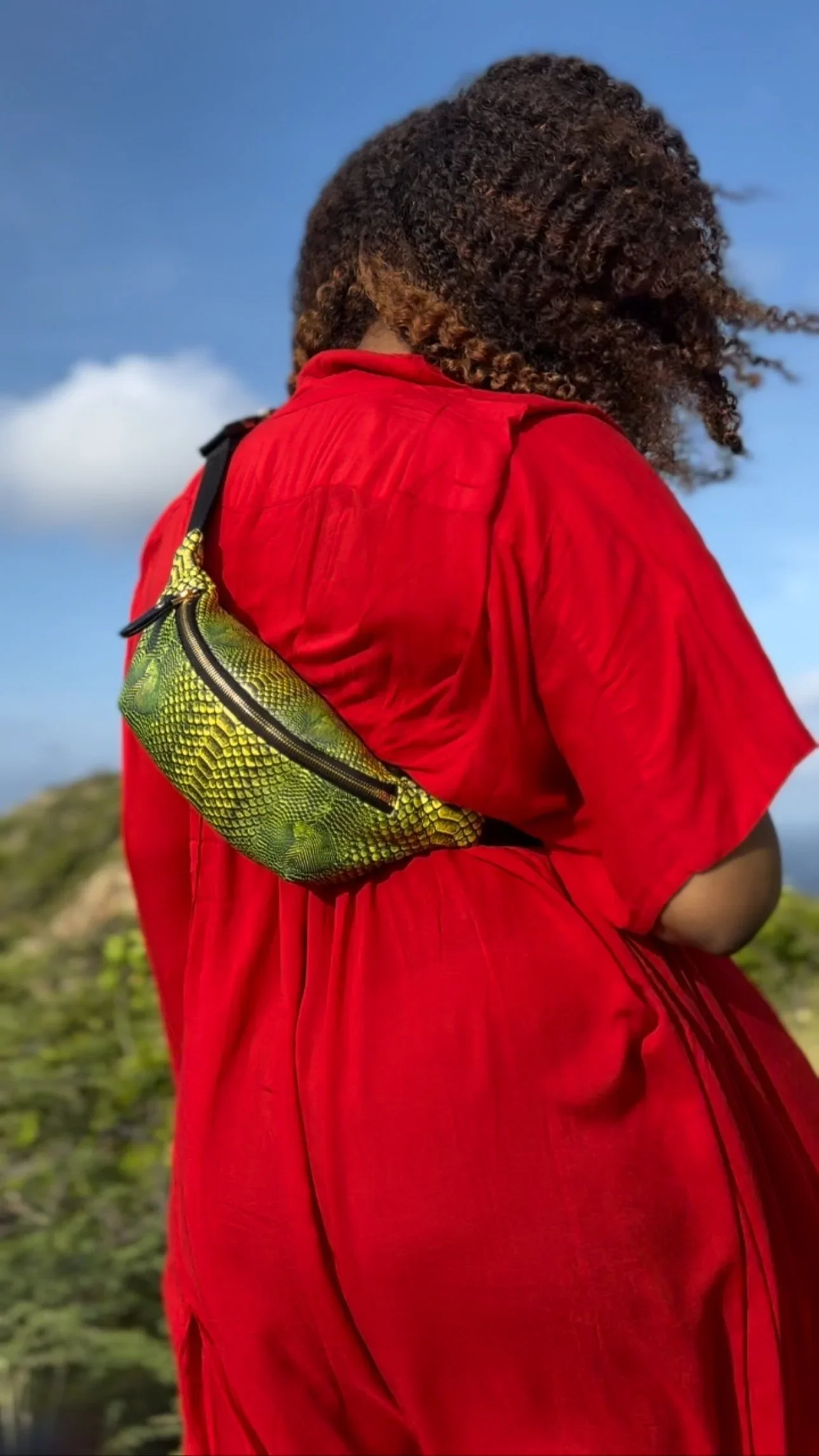A close-up photo of a model with curly hair, wearing a red jumpsuit with a textured bright green Fanny Pack across her body, against a bright blue sky with white clouds.
