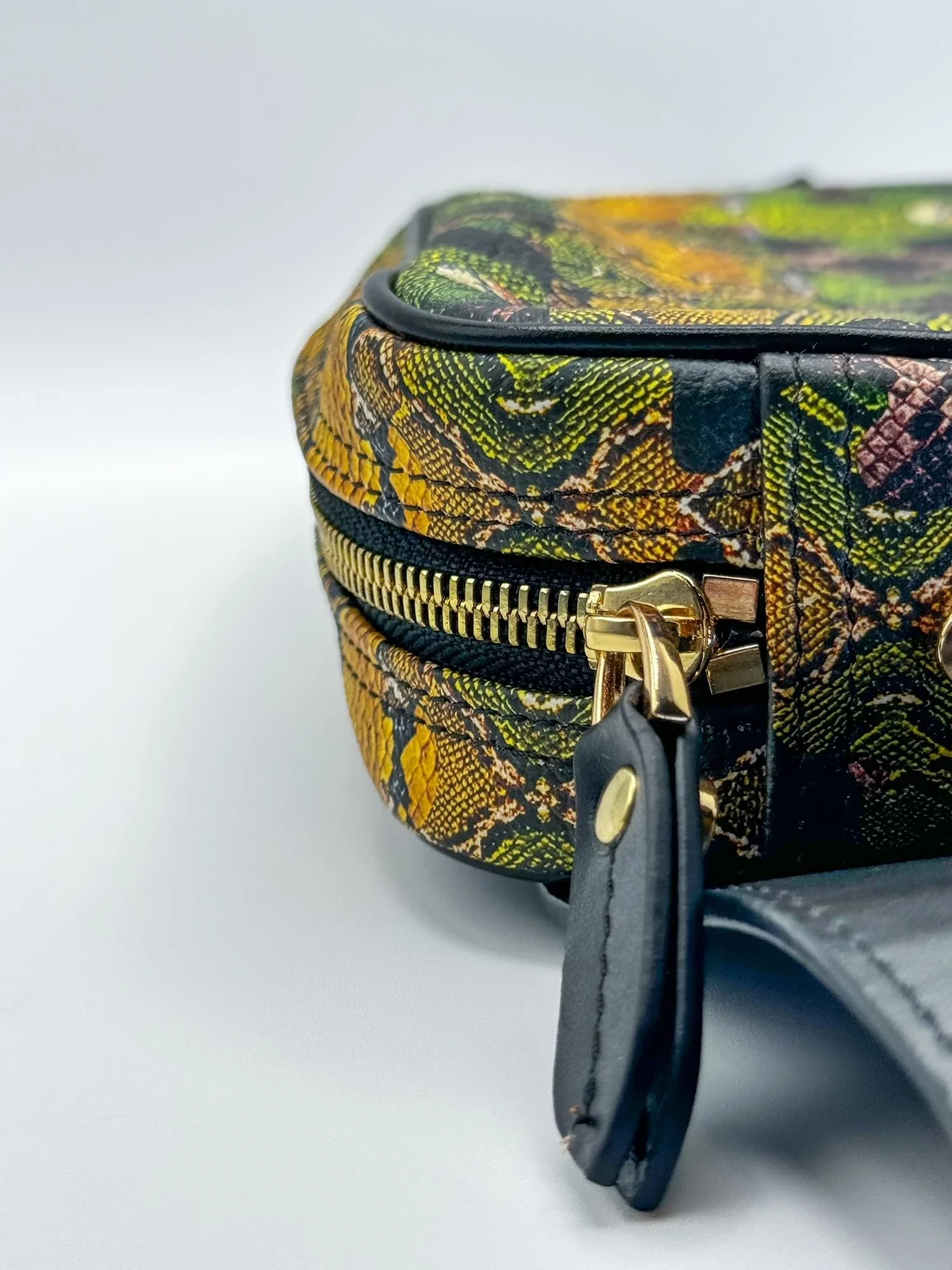 A close up photo of a multicolored Leather bag with a black waist strap, a black leather zipper pull and gold zipper. The background is white-grey.