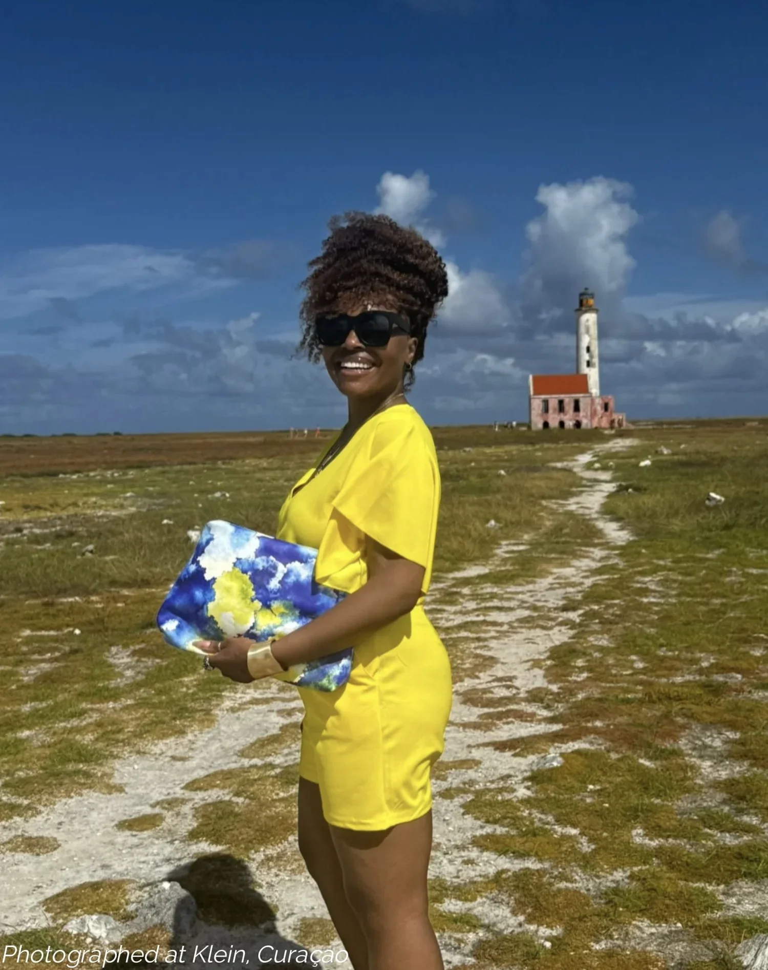 A model wearing a yellow shirt and shirt holding a large yellow, blue and white leather clutch in front of the iconic pink lighthouse on Klein Curaçao.
