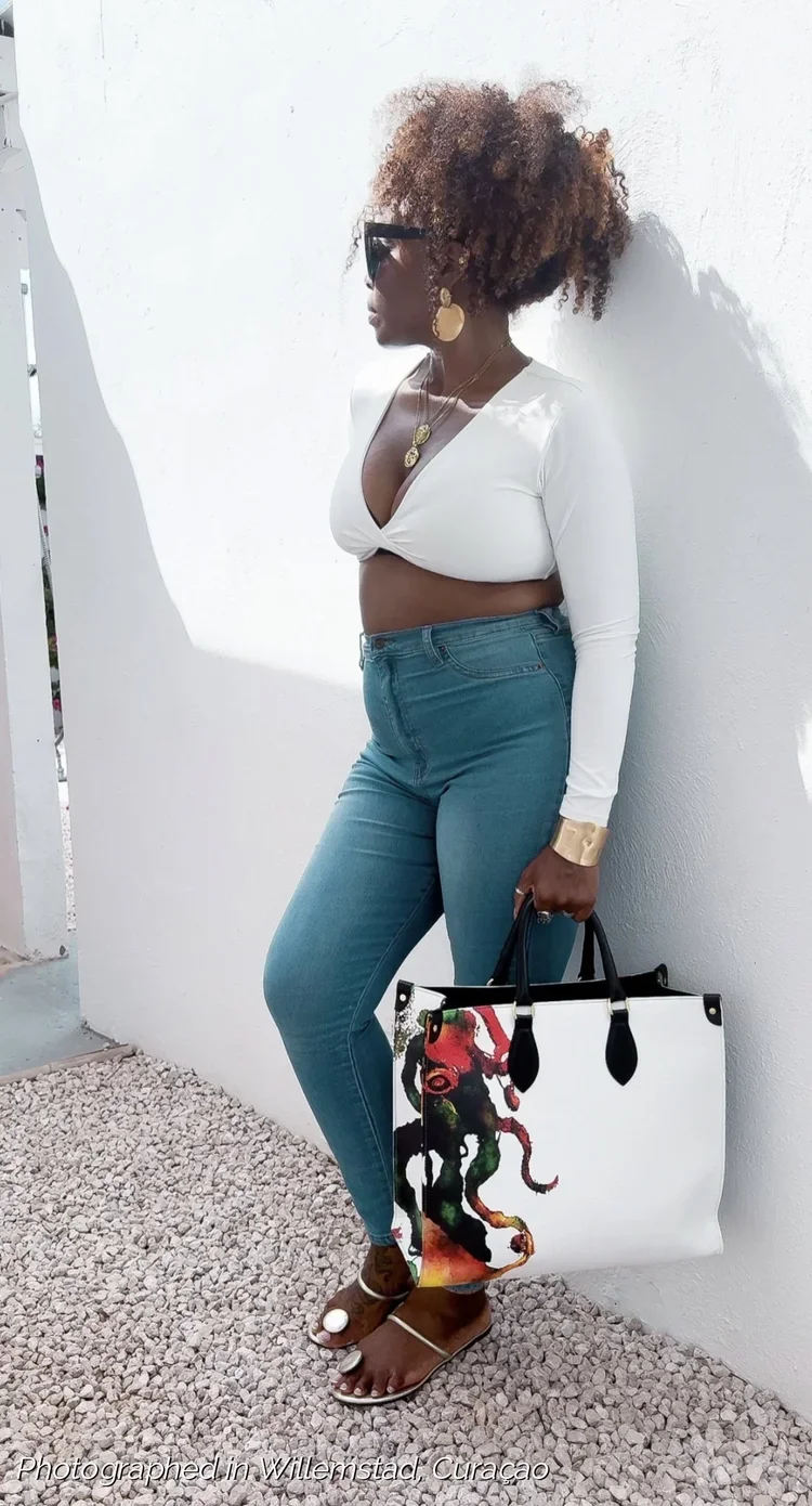 A model holding a large white and multicolored leather shopper bag, with a black leather handle. The model is standing outside with a white wall behind her, wearing a white cropped shirt and blue jeans.