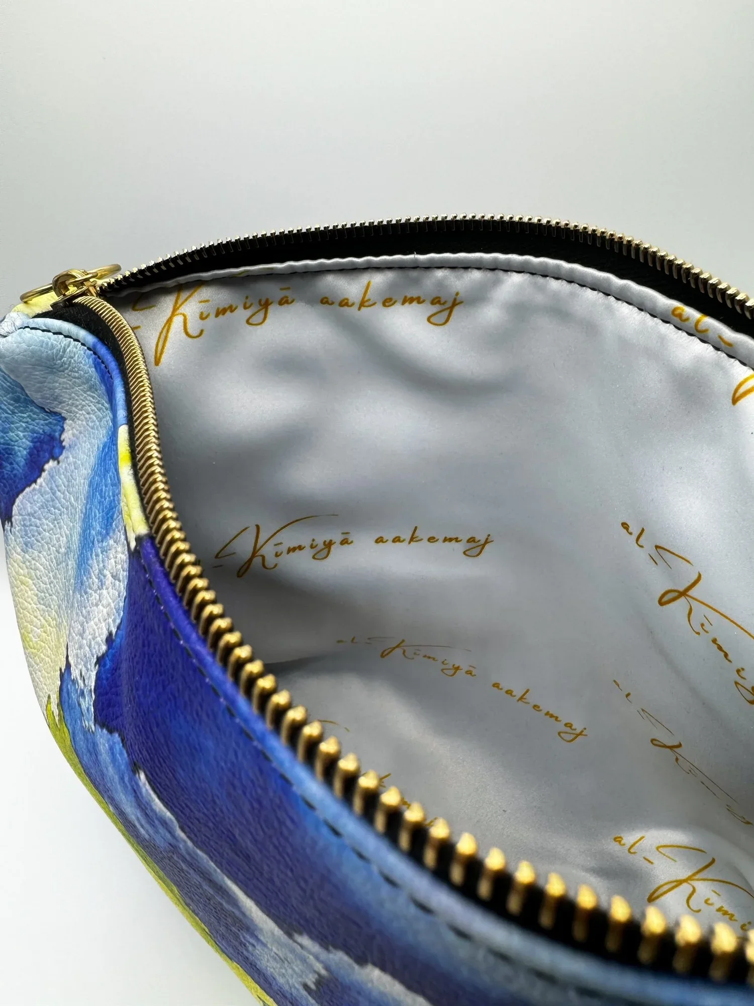 A close up photo of the white and gold satin interior lining of a large yellow, blue and white leather clutch with a gold circle pull and gold zipper, on a white background.