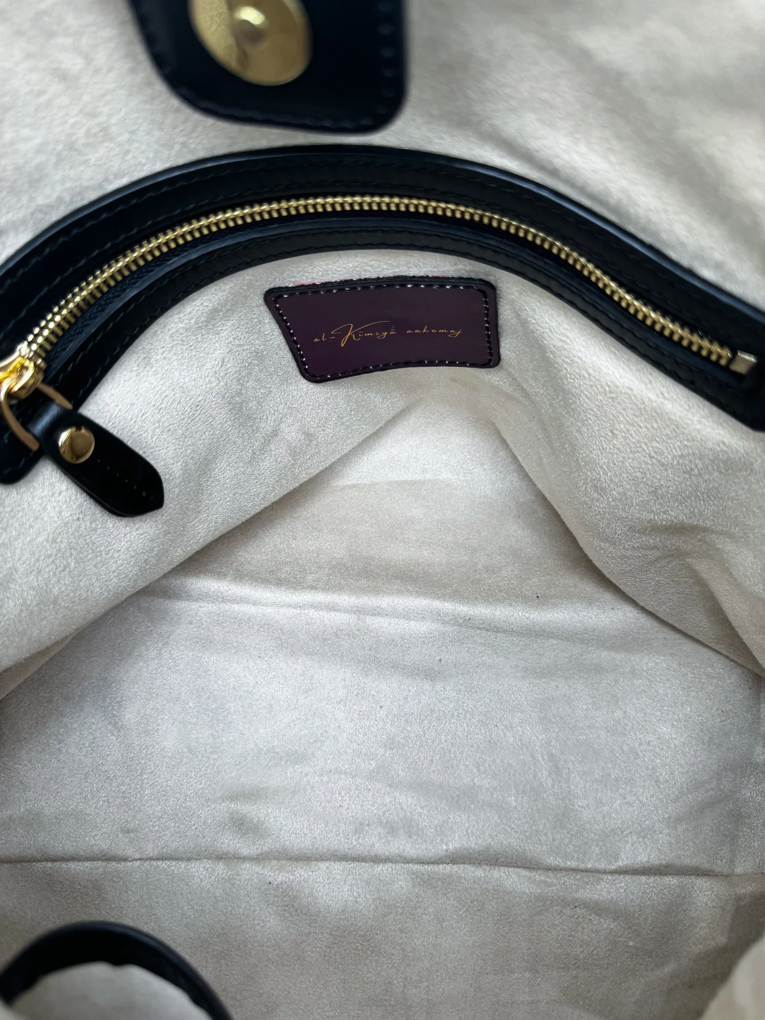 Close-up image of the beige suede interior of a leather handbag, featuring a gold and black leather zipper, gold magnetic lock button and burgundy interior label.