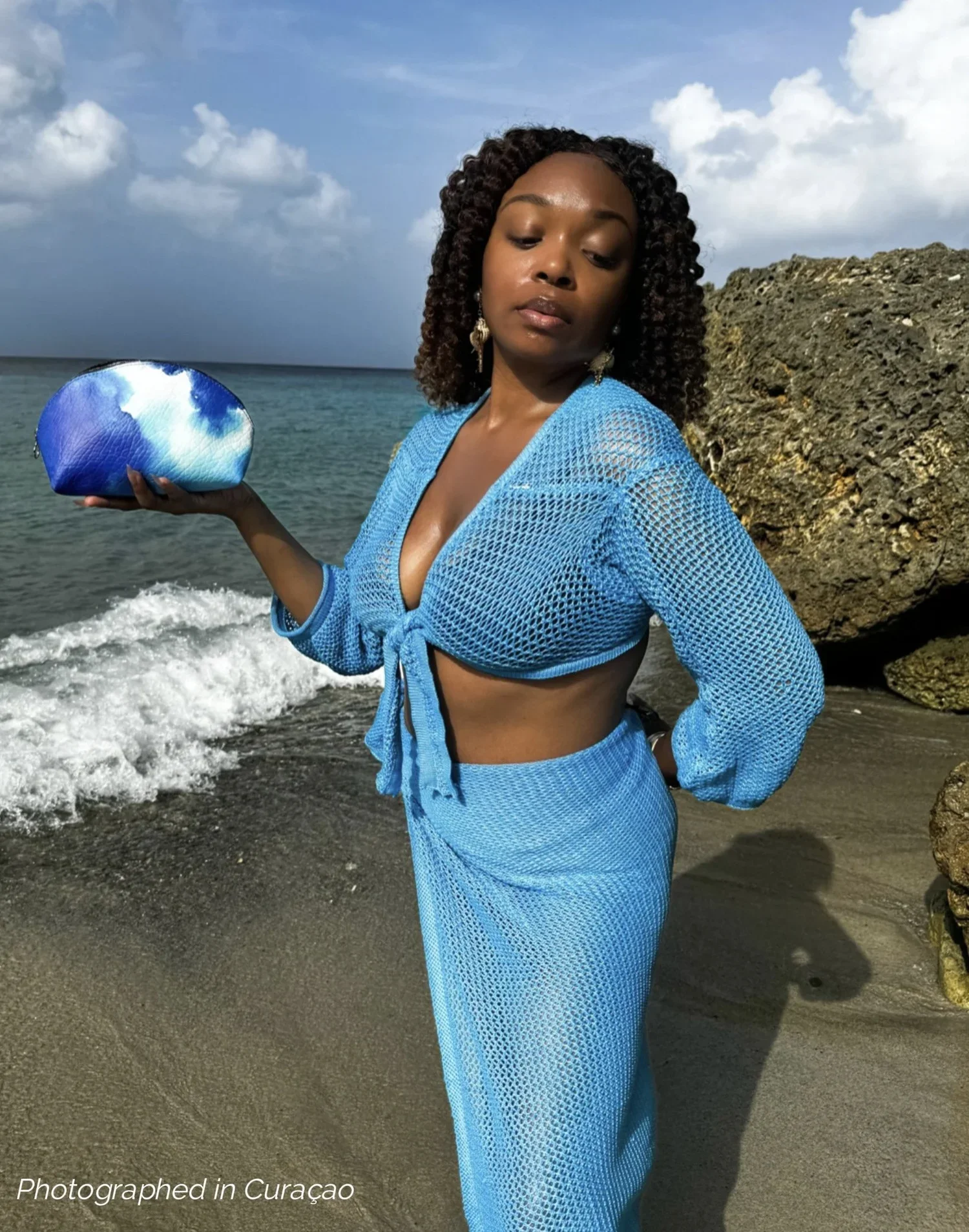 A model standing on a beach holding a blue multicolored leather shell coin purse, wearing a blue crochet knit two-piece set.
