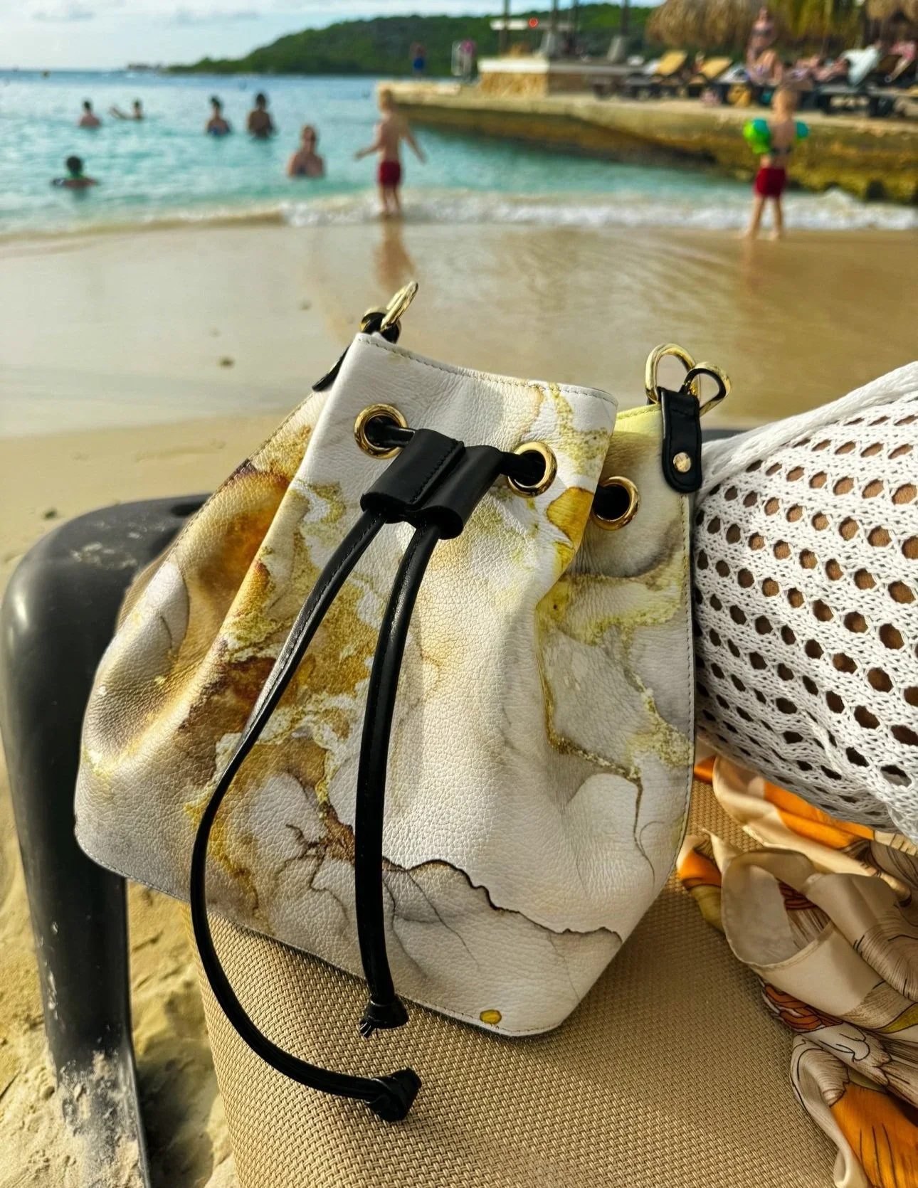 A  photo of a gold and white marble-patterned bucket bag with a black drawstring closure. The background of the photo is on a beach with blurry human figures swimming in the background,