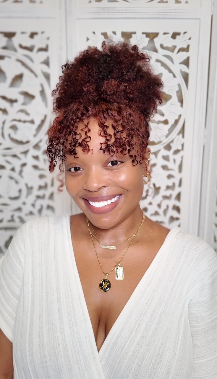 A model wearing a white shirt, wearing gold necklaces and earrings with copper-auburn colored curly hair. There is a white decorative backdrop in the background.