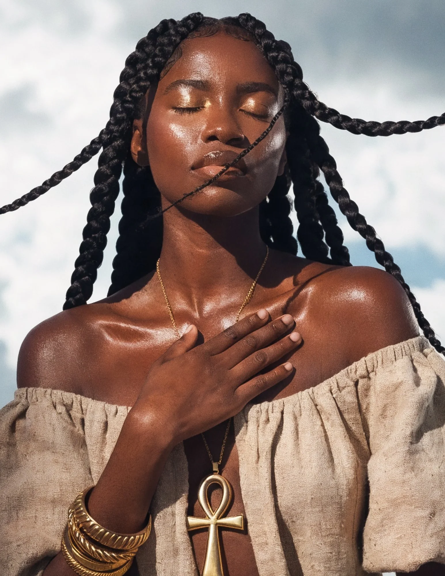 A young brown skinned woman stands in the sun, her eyes closed, her braids flowing into the wind. Her right hand is lying on her chest in a state of gratitude.