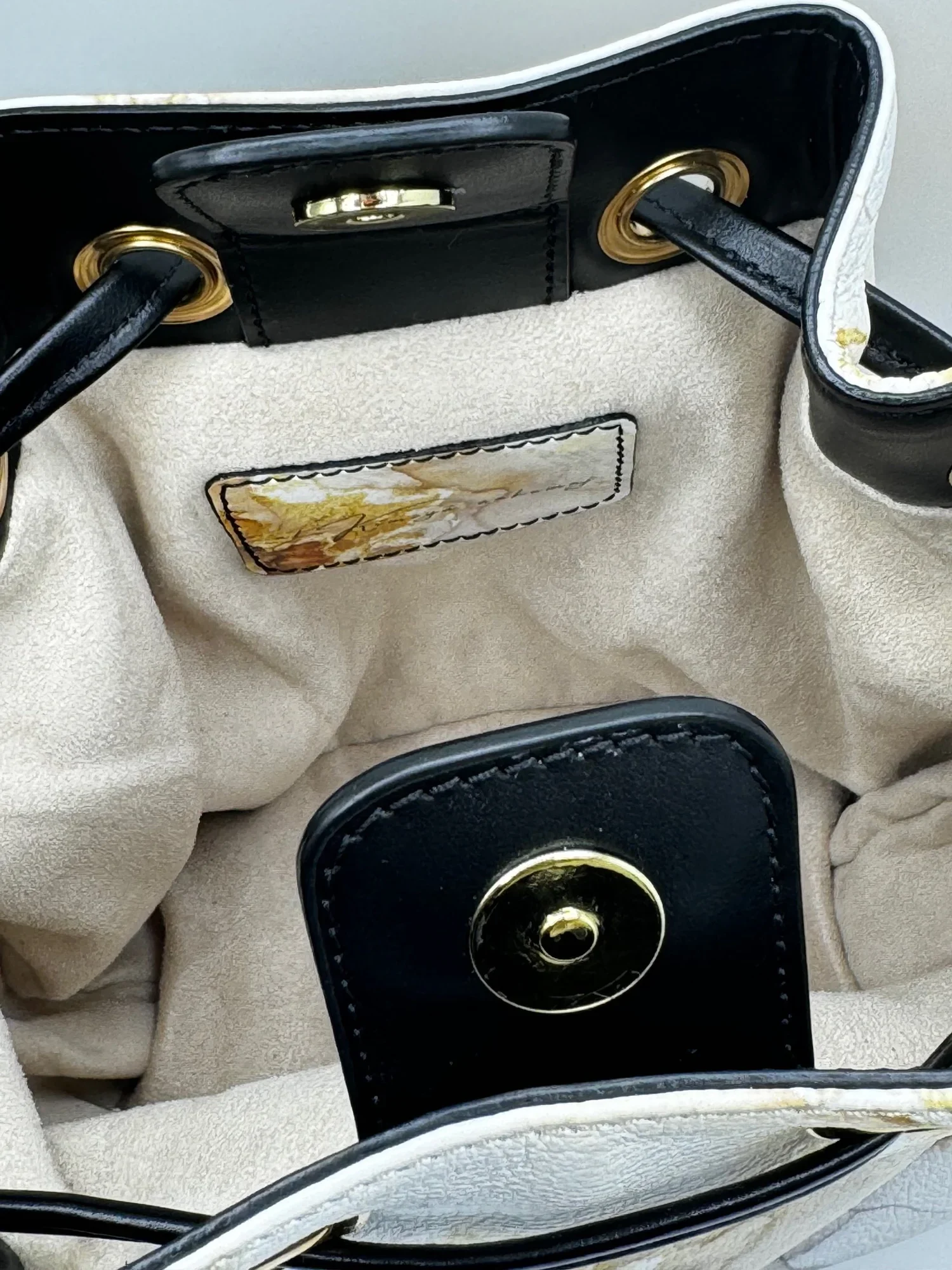 A close-up photo of the beige suede interior lining of a gold and white marble-patterned bucket bag with a black drawstring closure. The interior label is gold and white Marbled color.