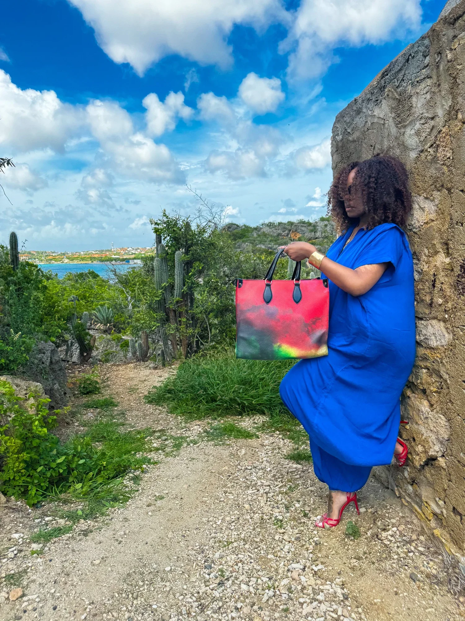 A photo of a model with curly hair standing in an ancient ruin site. She is wearing a blue jumpsuit, holding a large red, black and green leather shopper bag.
