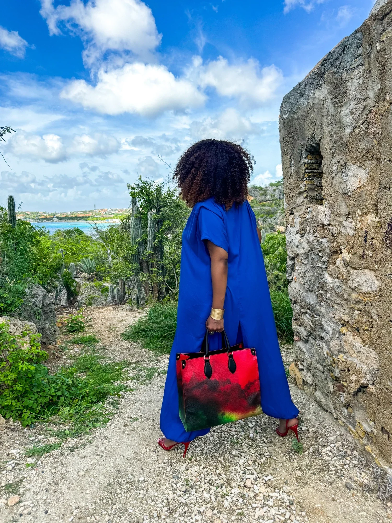 A photo of a model with curly hair standing in an ancient ruin site. She is wearing a blue jumpsuit, holding a large red, black and green leather shopper bag.