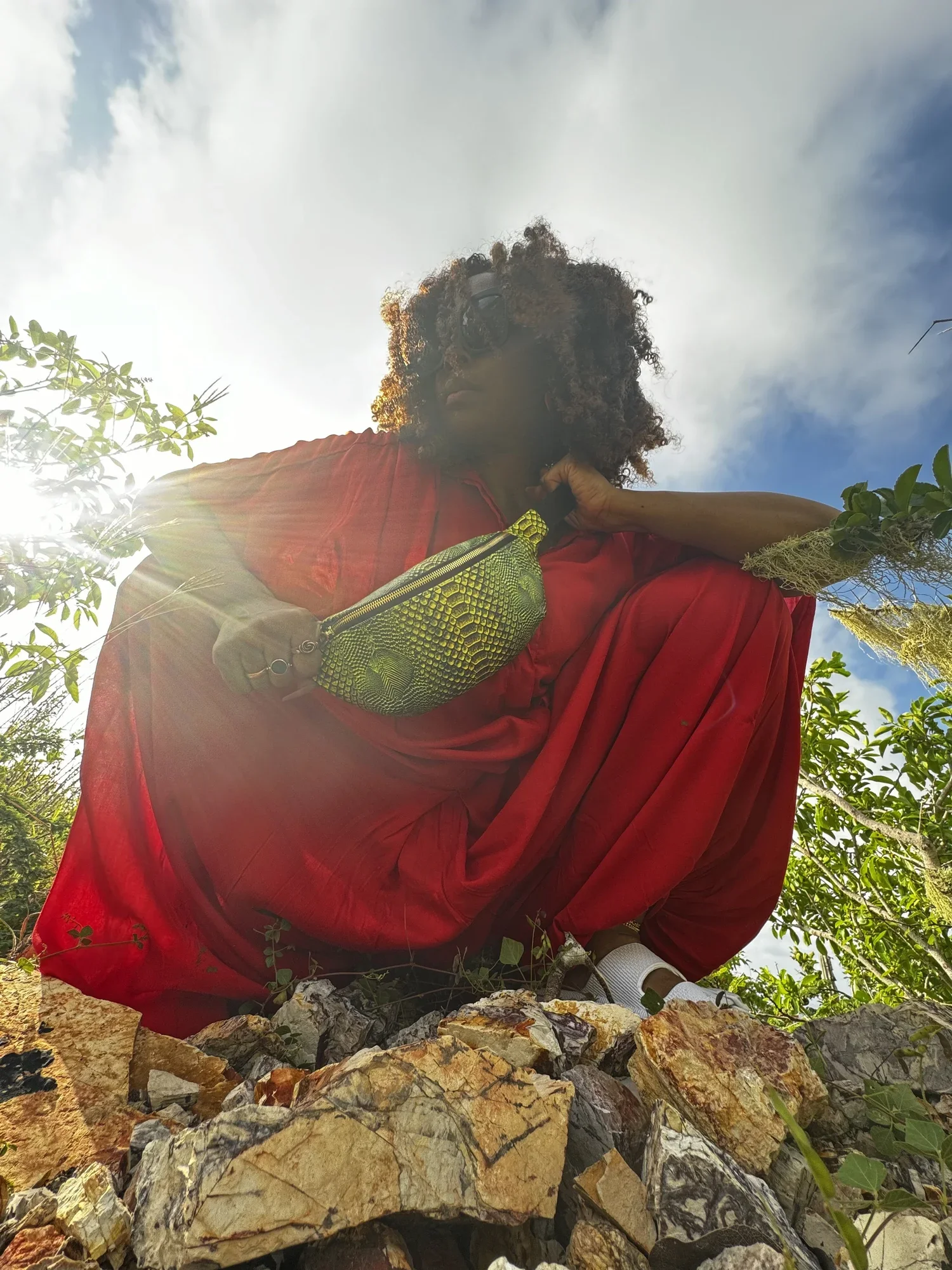 A model with curly hair, wearing a red jumpsuit with a textured bright green Fanny Pack across her body, against a bright blue sky with white clouds.
