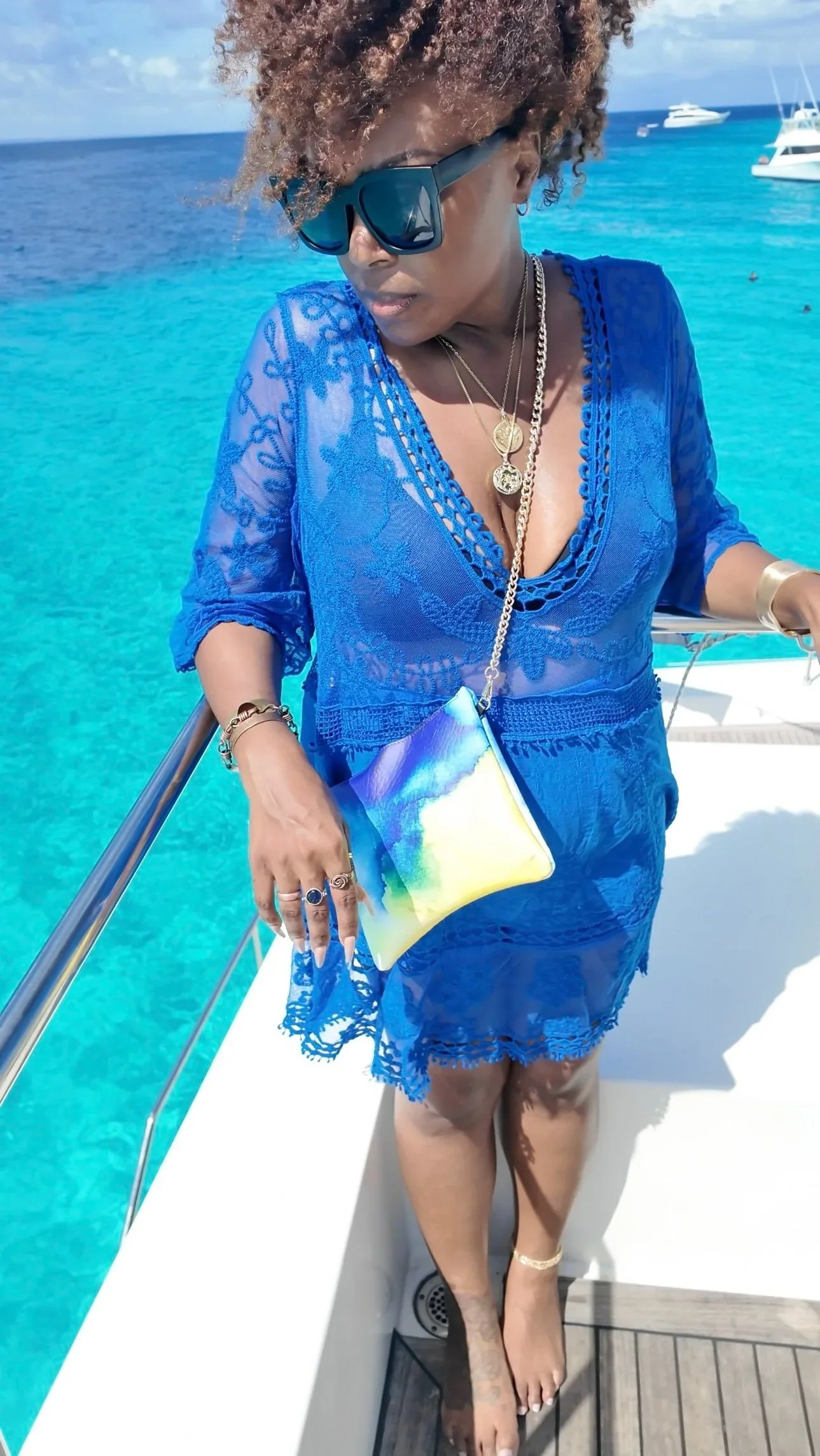 A model with curly hair wearing a blue dress, standing on a yacht looking at the blue ocean behind her, wearing a yellow and blue crossbody purse, with a gold chain.